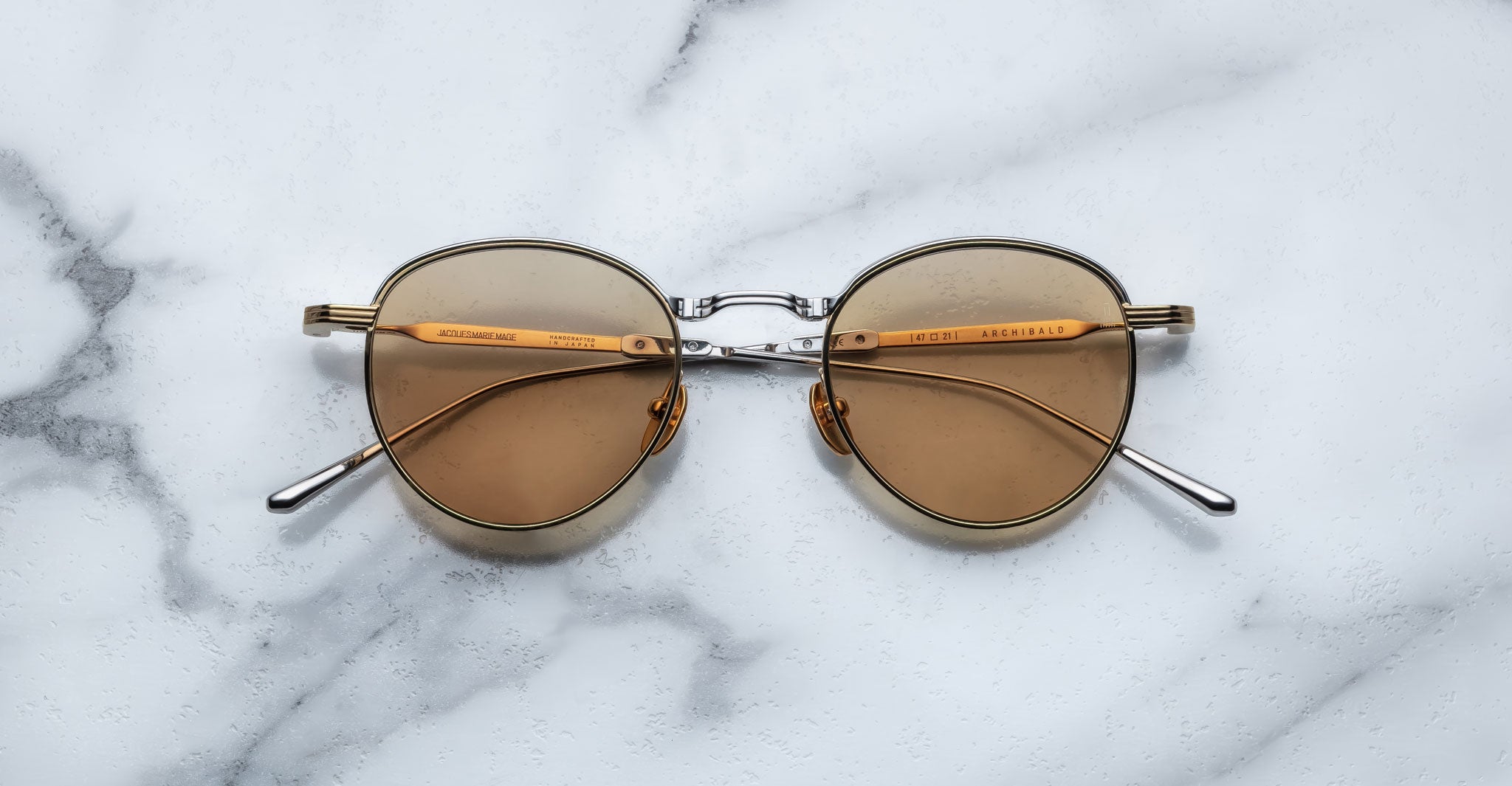 A pair of round, gold-framed sunglasses with brown lenses is placed on a white marble surface. The arms of the sunglasses are metallic with visible branding on the inner side of the arms.