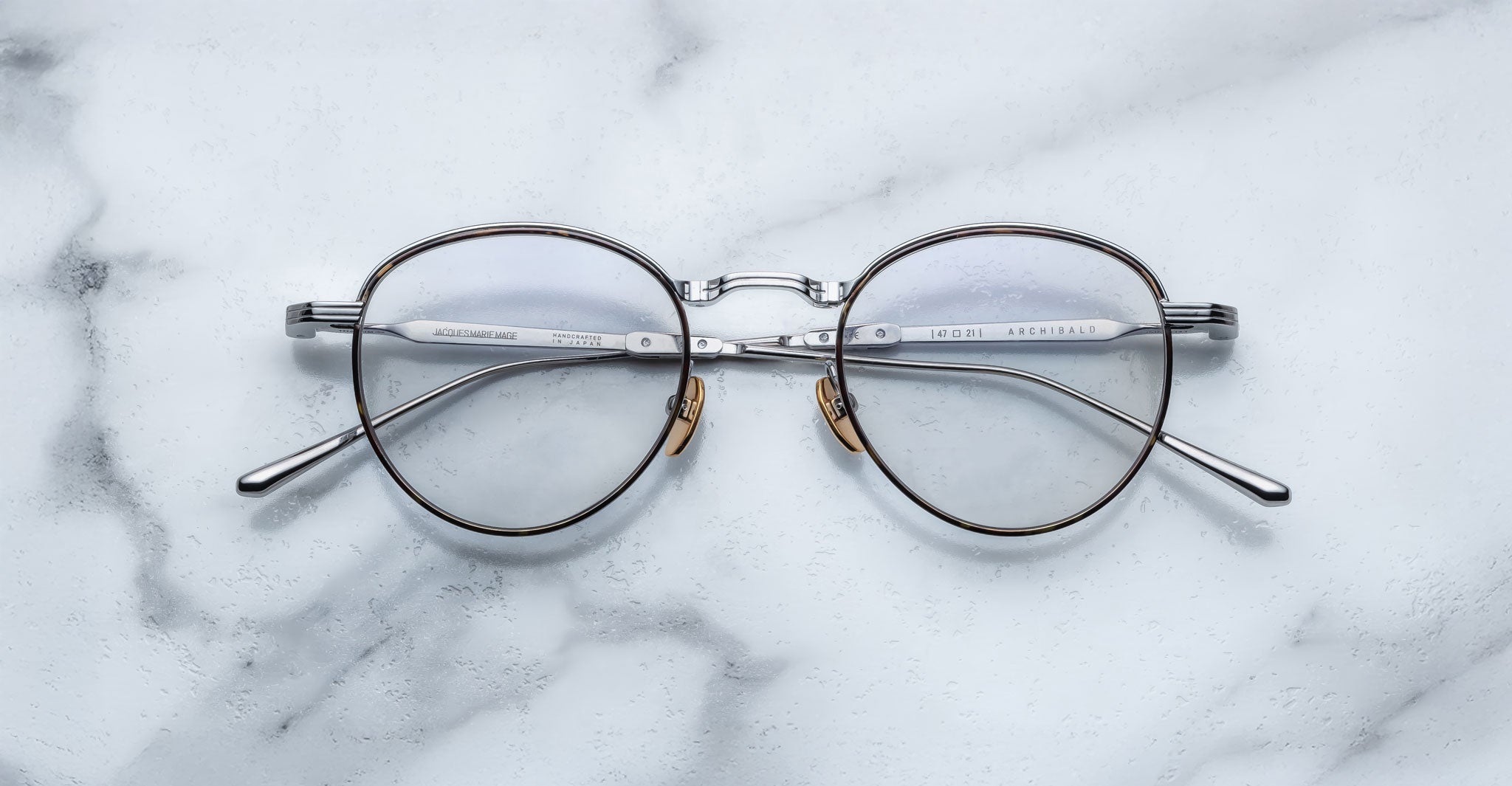A pair of Jacques Marie Mage Archibald round, silver-framed optical frame with thin metal arms rests on a white marble surface with grey veining.