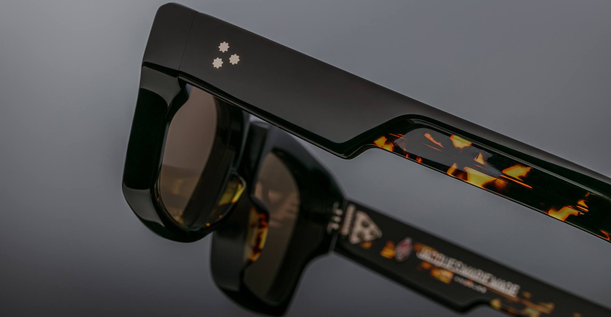 Close-up of Jacques Marie Mage Ashcroft sunglasses with thick black and tortoiseshell-patterned arms and two small white star accents on the frame, reflected on a glossy surface.