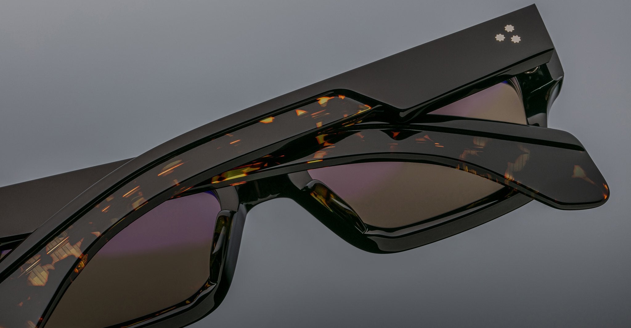 Close-up of two Jacques Marie Mage Ashcroft sunglasses: one with a glossy black frame and another in tortoiseshell, stacked together against a neutral background.