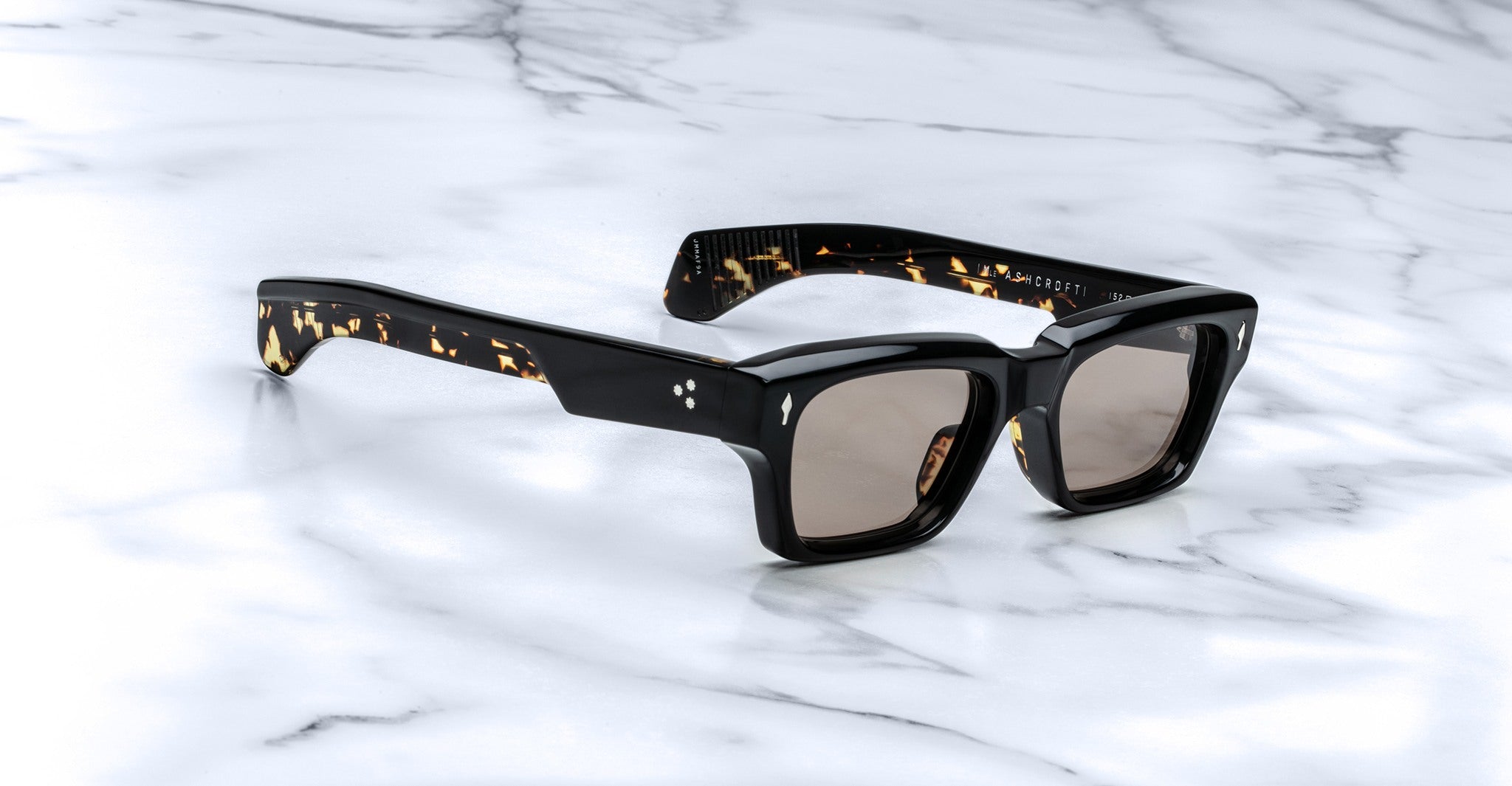 The Jacques Marie Mage Ashcroft, black thick-framed sunglasses with tinted lenses and tortoiseshell accents, rests on a white marble surface.