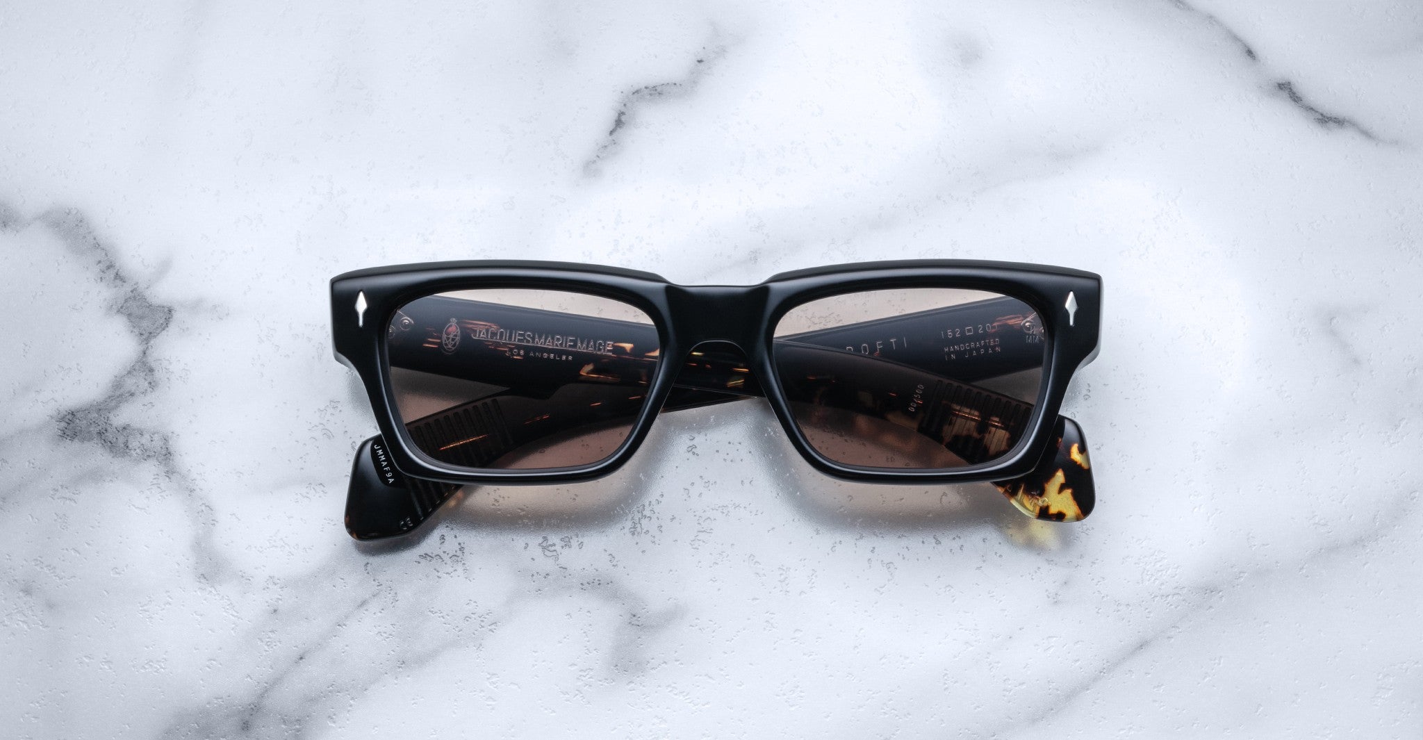 The Jacques Marie Mage Ashcroft black rectangular sunglasses with dark lenses rest on a white marble surface, their lenses reflecting a blurred indoor scene with lights and text.