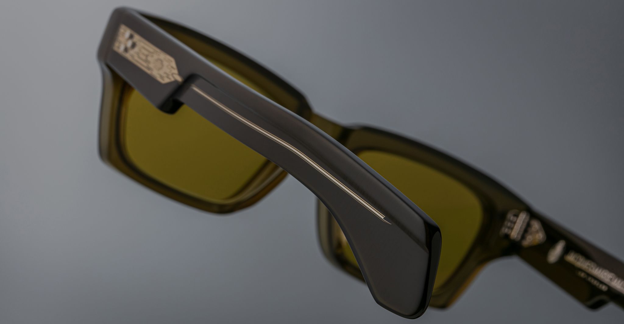 Close-up of Jacques Marie Mage Ashcroft sunglasses—dark, rectangular frames with green-tinted lenses and metal temple accents—set against a plain gray background.