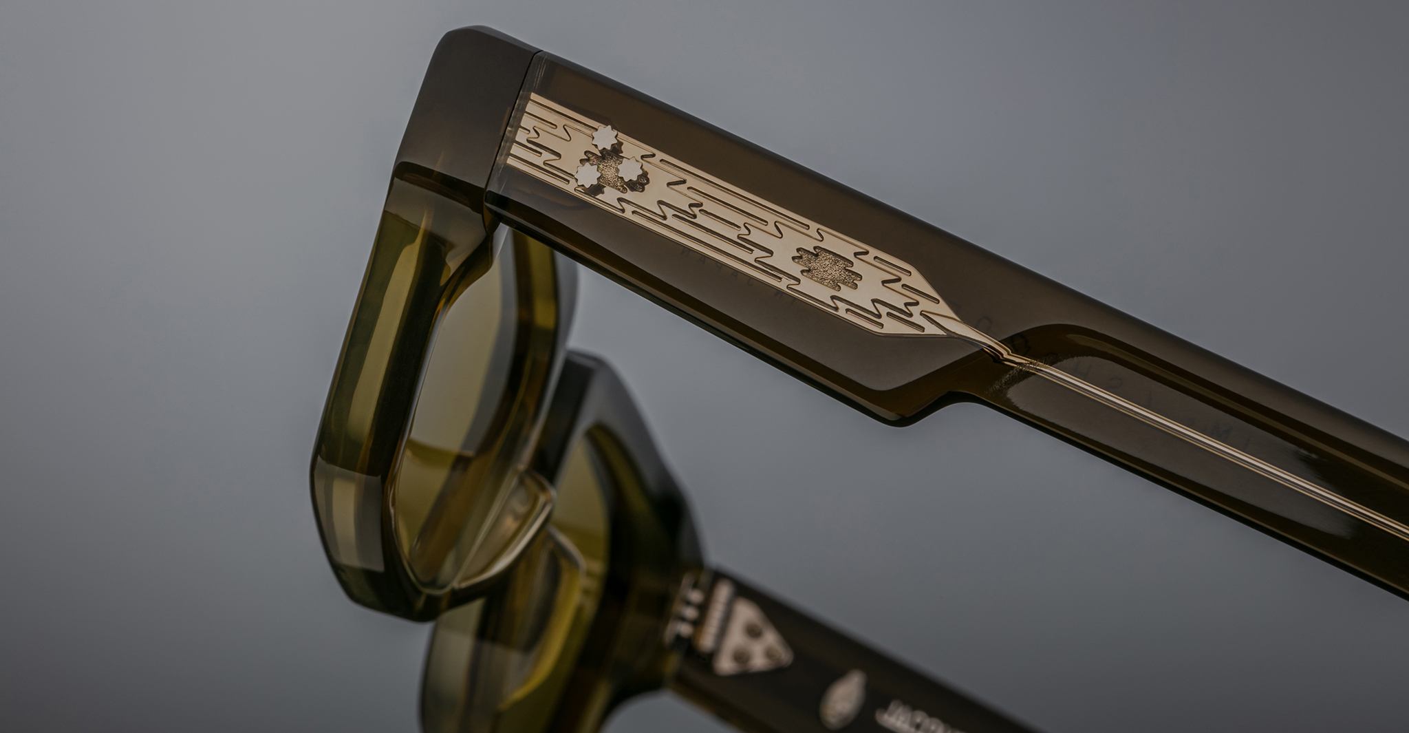 A close-up of the Jacques Marie Mage Ashcroft eyeglasses arm in translucent olive green, showcasing a metallic inlay engraved with lightning bolts and stars, with the glasses’ reflection visible on the surface below.