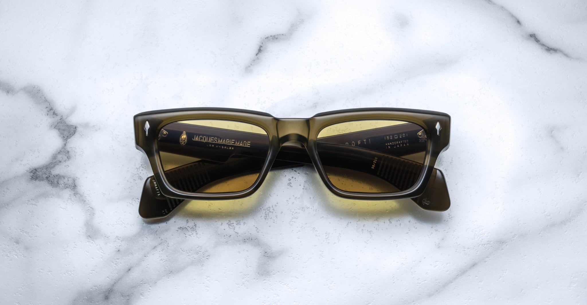 The Jacques Marie Mage Ashcroft sunglasses, featuring rectangular yellow-tinted lenses and dark frames, are displayed face up on a white marble surface.