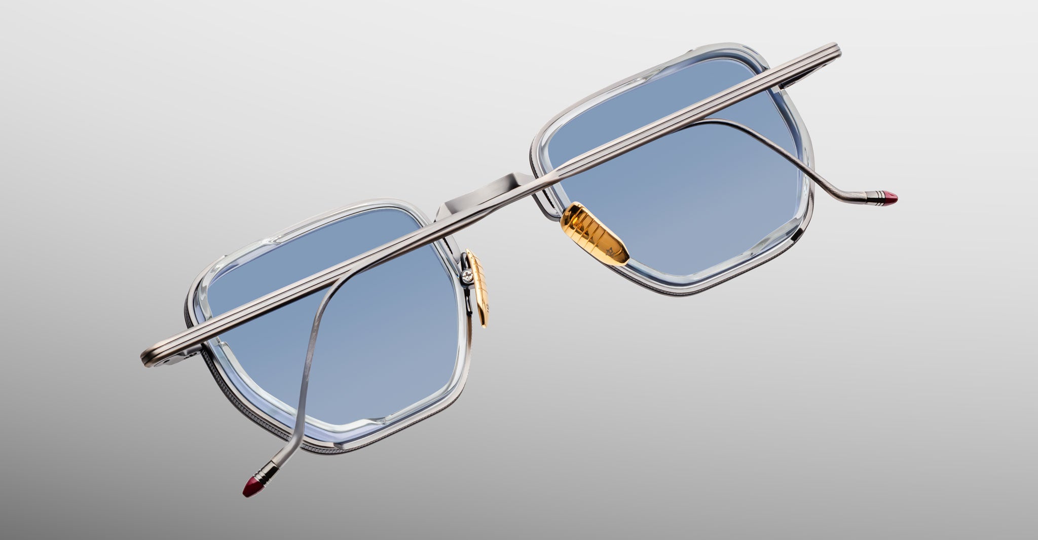 A pair of Jacques Marie Mage Atkins sunglasses with silver metal frames, blue square lenses, gold nose pads, and red-tipped arms displayed on a light gray background—luxurious handcrafted Japanese eyewear.