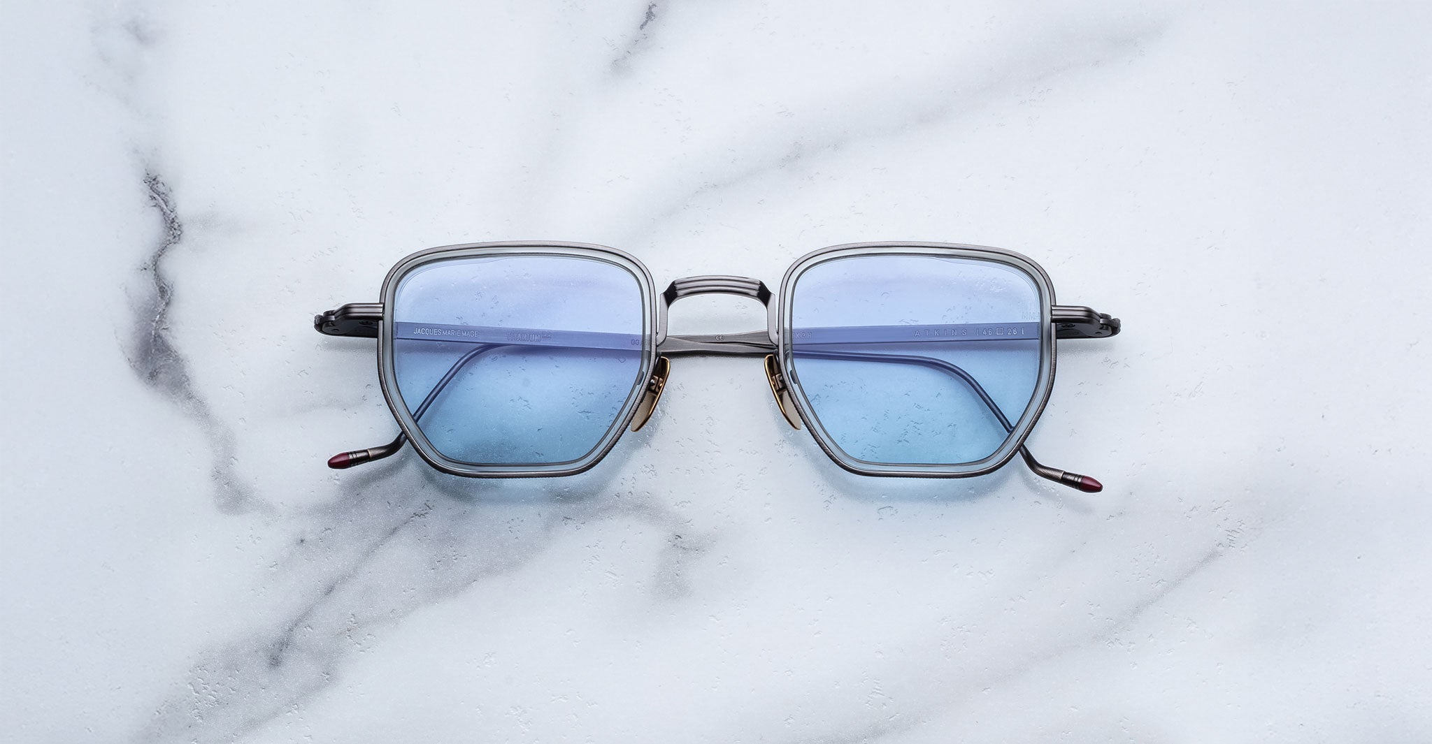 Jacques Marie Mage Atkins eyeglasses from the Titanium Series, with square blue-tinted lenses and slim metal frames, rest on a white marble surface veined in gray.