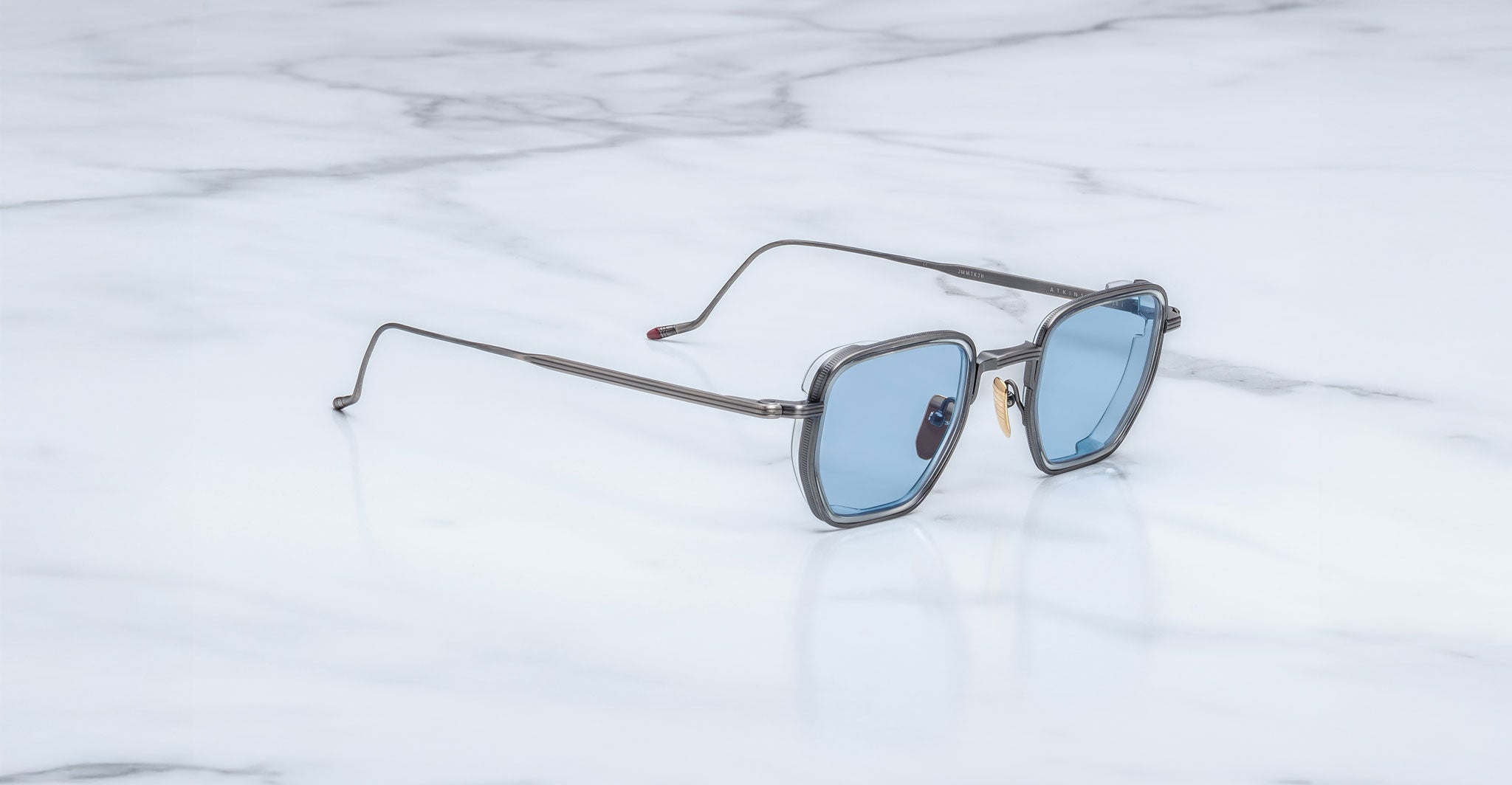 The Jacques Marie Mage Atkins, modern square-shaped sunglasses from the Titanium Series with thin metal frames and blue-tinted lenses, rest elegantly on a white marble surface.