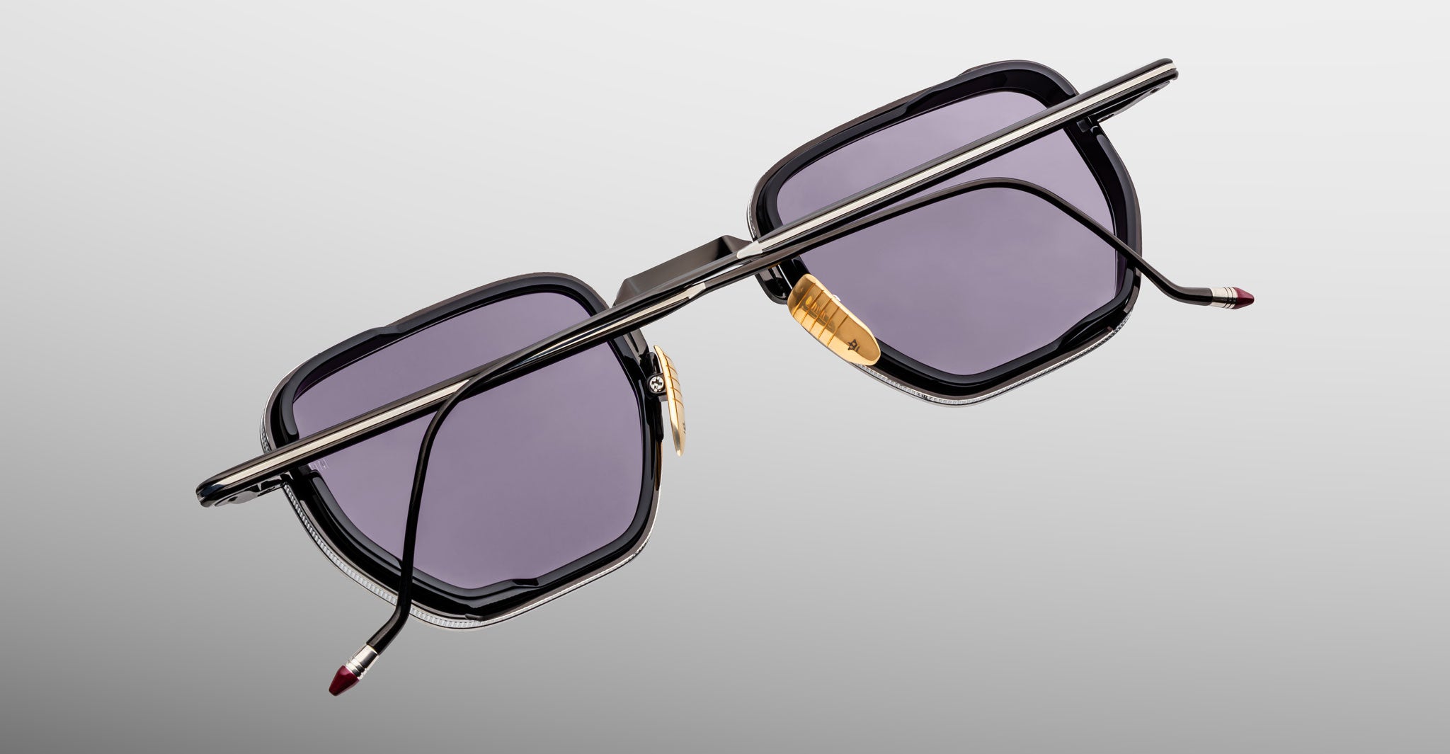 A pair of Jacques Marie Mage Atkins sunglasses featuring a rectangular black frame, purple-tinted lenses, gold nose pads, and thin metal arms is displayed upside down on a light gradient background.