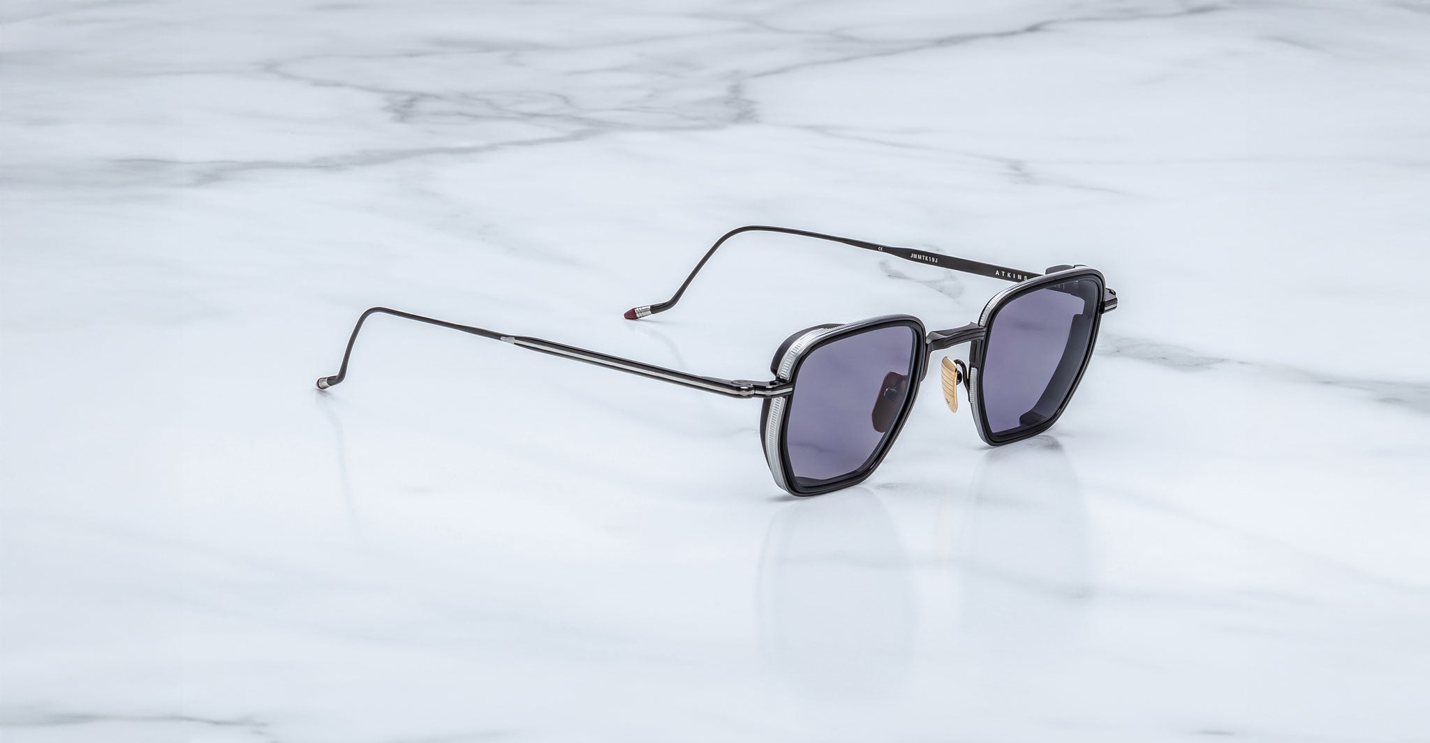 A pair of Jacques Marie Mage Atkins sunglasses with black square frames and dark lenses rests on a white marble surface.