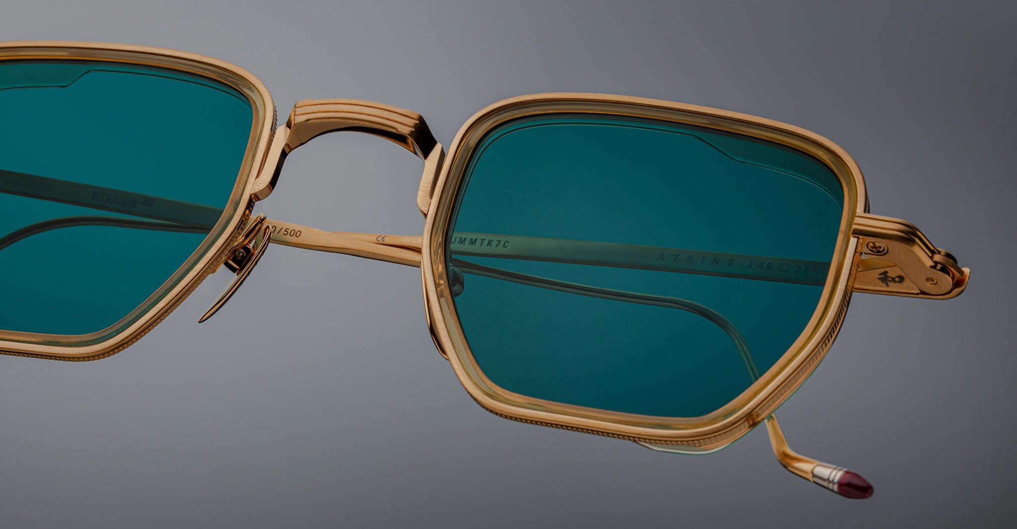 Close-up of Jacques Marie Mage Atkins sunglasses, square gold rims with teal lenses, viewed from the front.