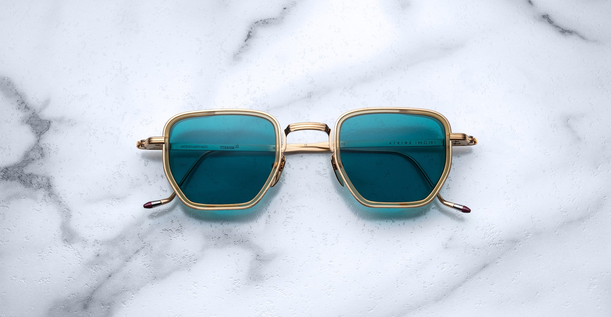 Jacques Marie Mage Atkins square sunglasses with gold frames and blue lenses, viewed from above.
