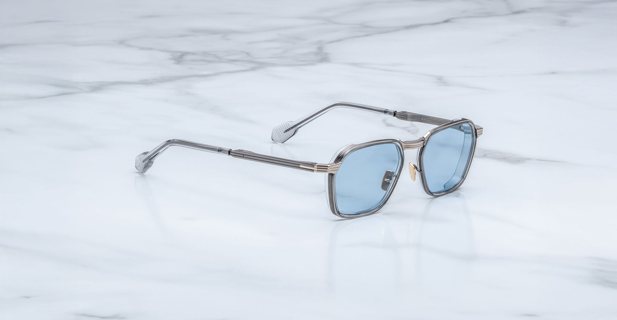 The Jacques Marie Mage Beckett sunglasses, handcrafted in Japan with a limited-edition frame, feature silver accents and blue-tinted lenses displayed elegantly on white marble.