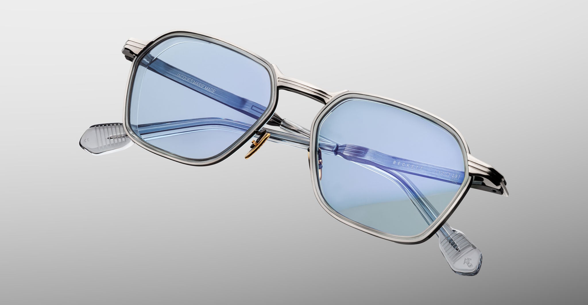 The Jacques Marie Mage Beckett eyeglasses feature a rectangular silver frame, light blue tinted lenses, and clear temple tips, handcrafted in Japan and displayed on a soft gradient background.