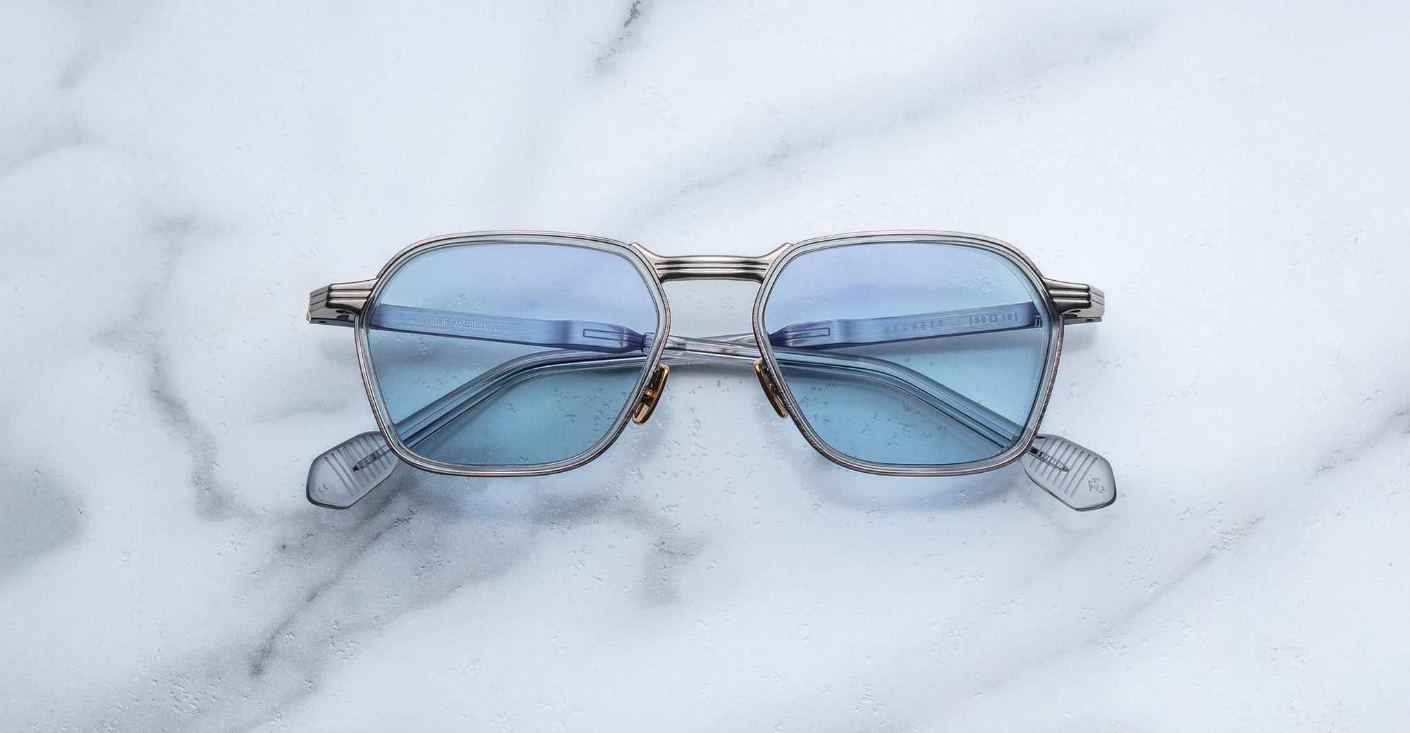 The Jacques Marie Mage Beckett features stylish, square-shaped silver frames and blue-tinted lenses, handcrafted in Japan, shown resting on a white marble surface.
