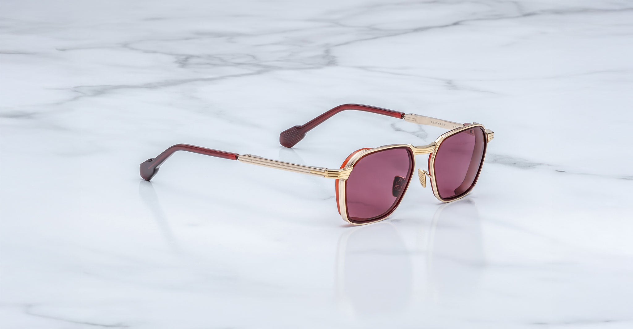A pair of limited-edition Jacques Marie Mage Beckett sunglasses with gold frames, maroon temples, and pink-tinted lenses, handcrafted in Japan, elegantly rests on a white marble surface.