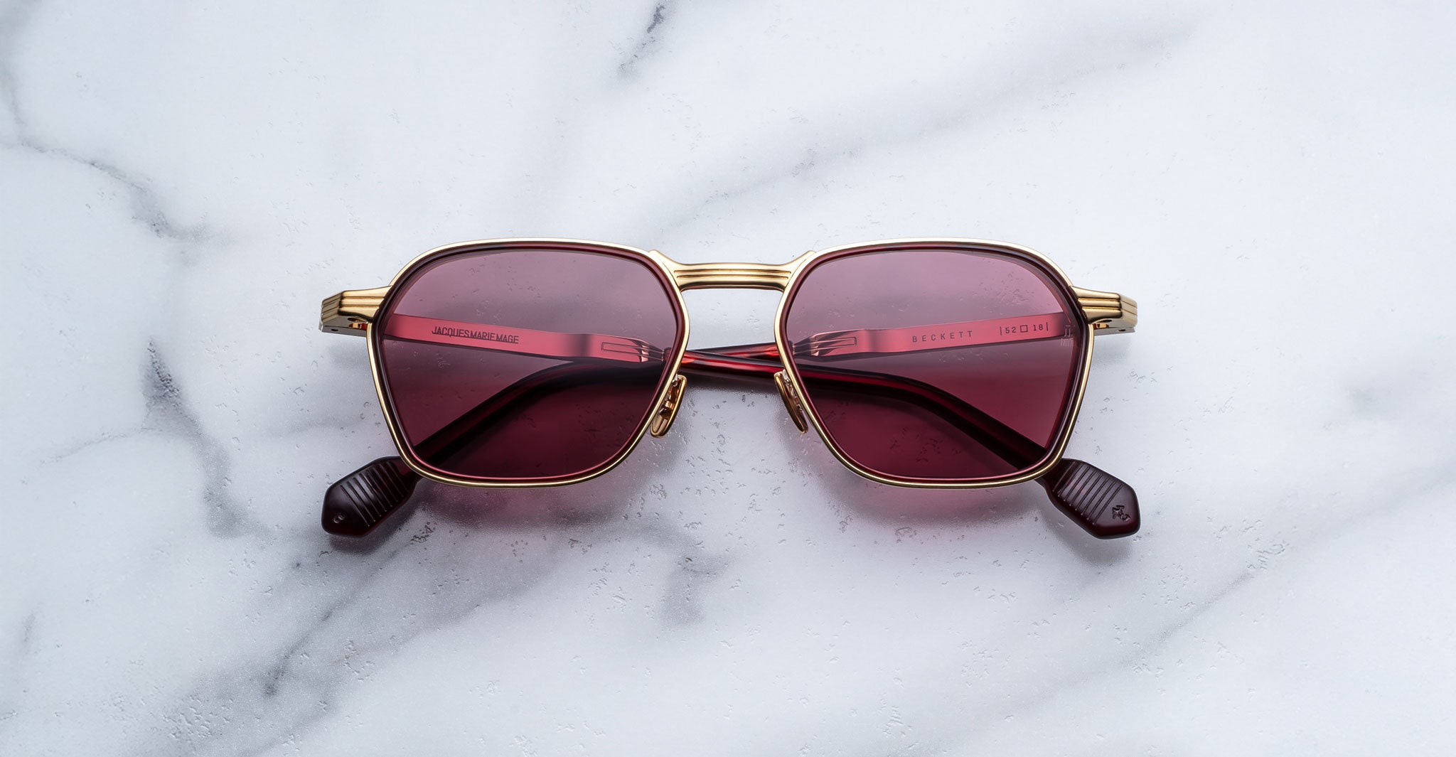 The Jacques Marie Mage Beckett is a limited-edition frame with gold rims and geometric dark red tinted lenses, handcrafted in Japan and displayed on a white marble surface.