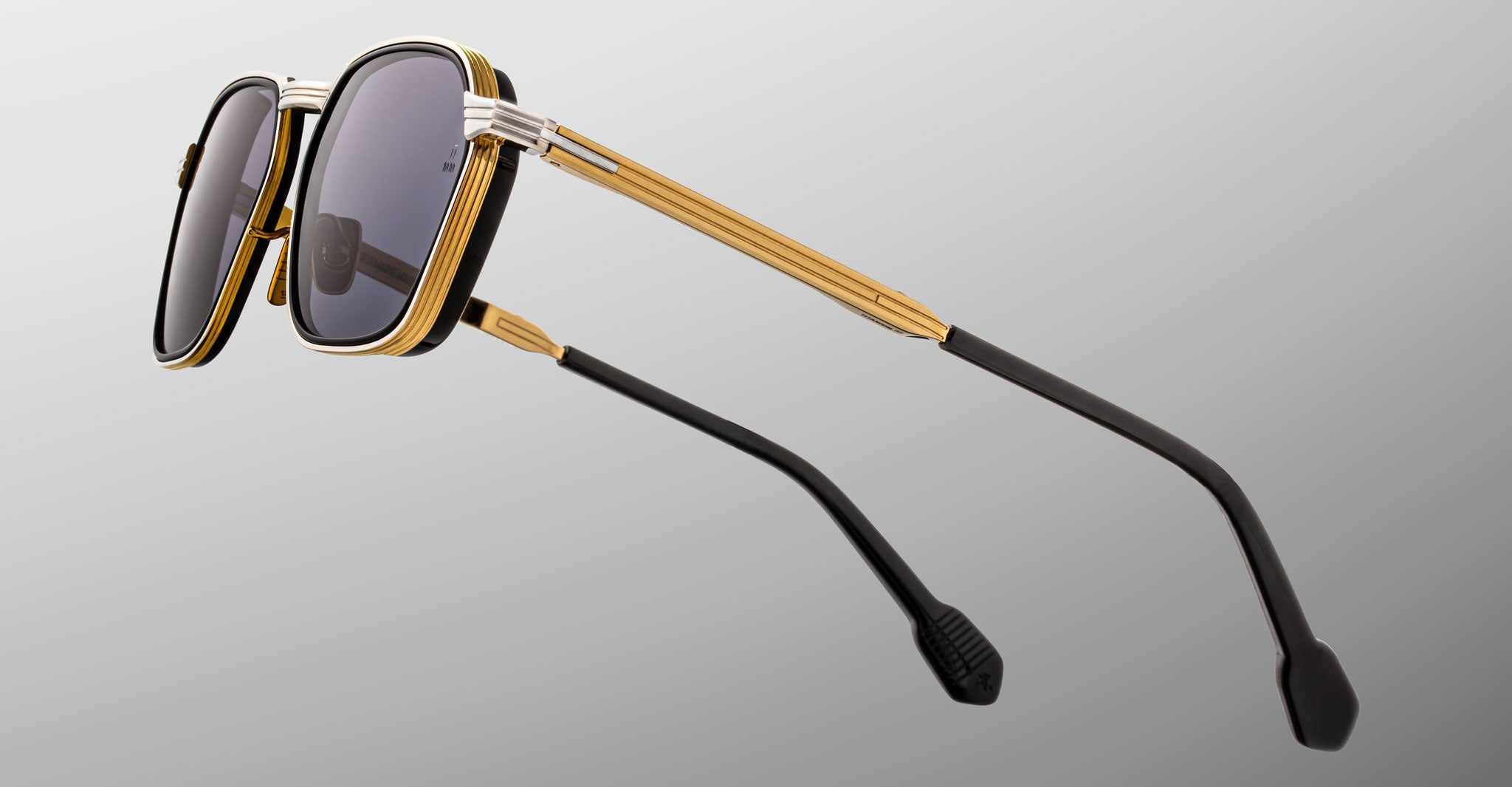 Close-up of the Jacques Marie Mage Beckett sunglasses, a limited-edition frame with gold and black accents, dark lenses, and black temple tips, handcrafted in Japan and set against a plain gray background.