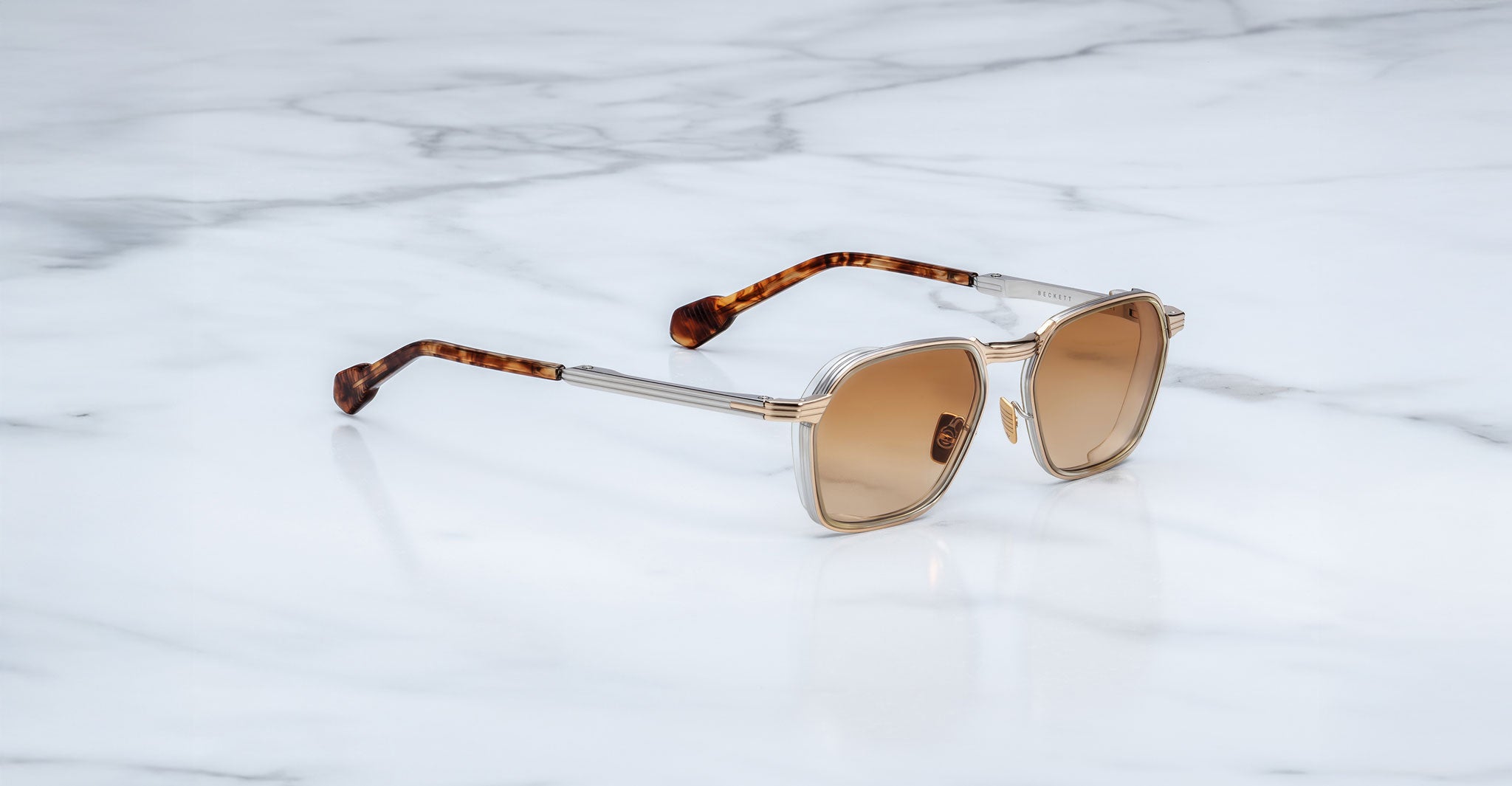 Jacques Marie Mage Beckett sunglasses, handcrafted in Japan with square frames, light brown lenses, and tortoiseshell-patterned arms, rest on a white marble surface.
