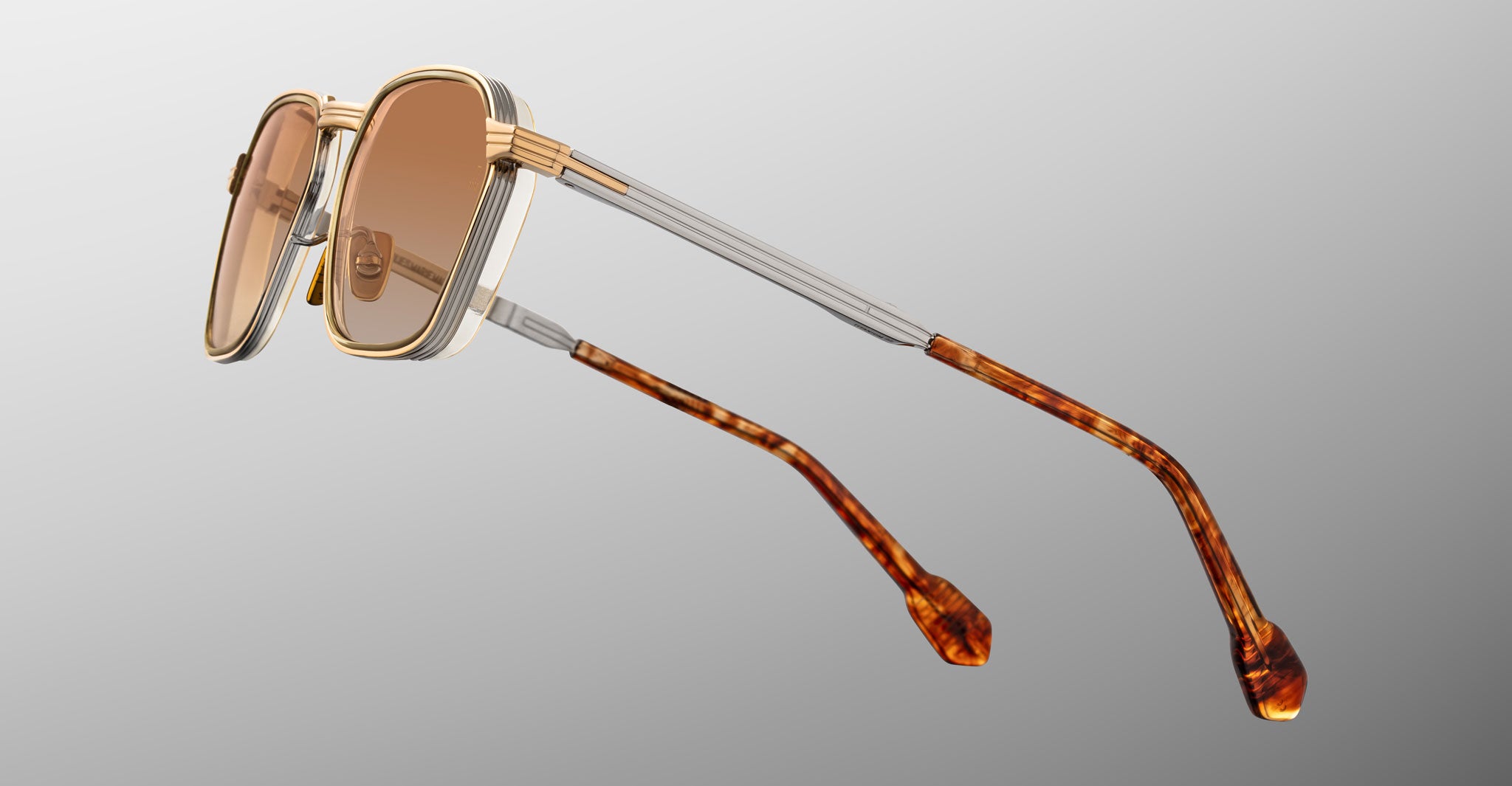 Jacques Marie Mage Beckett sunglasses with gold and silver frames, brown-tinted lenses, and tortoiseshell-patterned arms handcrafted in Japan are displayed against a gradient gray background.