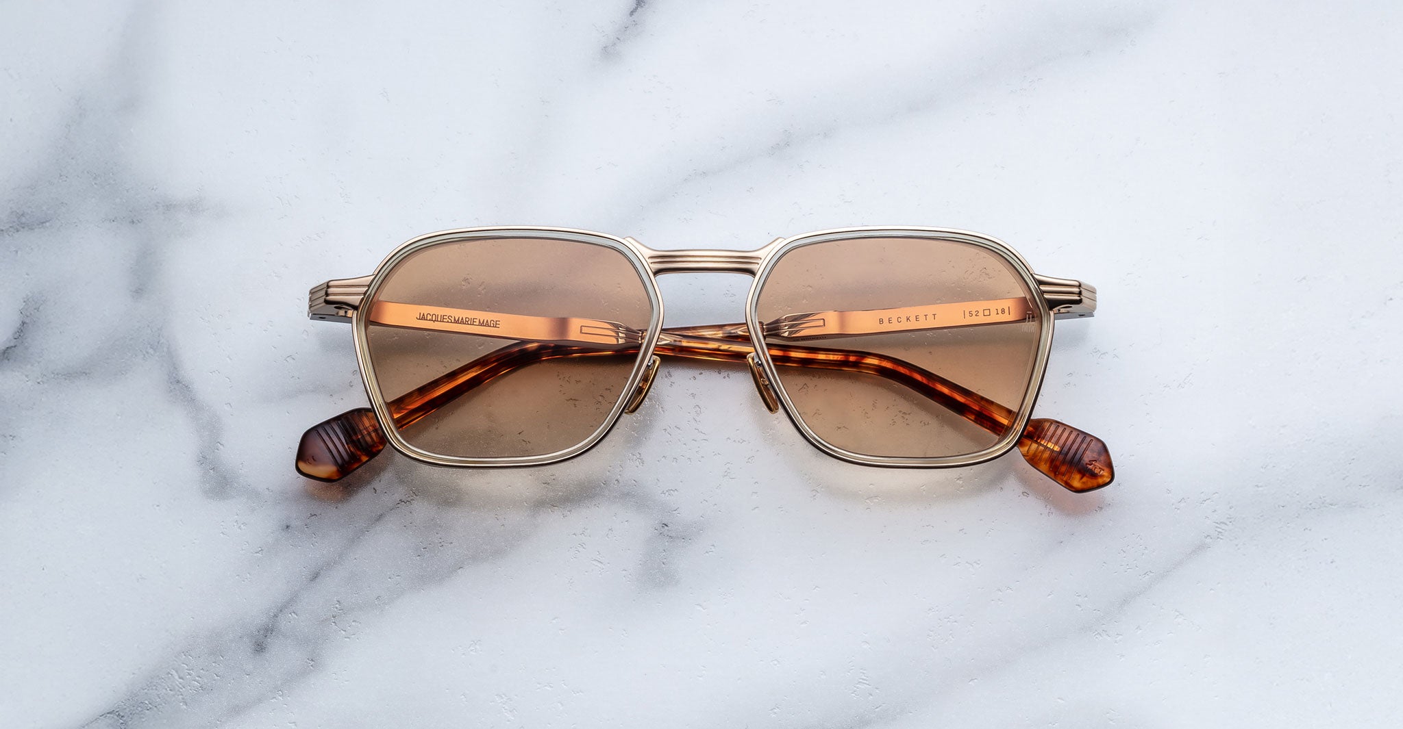 A pair of limited-edition Jacques Marie Mage Beckett sunglasses with gold rims and brown-tinted, square lenses rests on a white marble surface. Handcrafted in Japan, they feature matching brown temples and tips.
