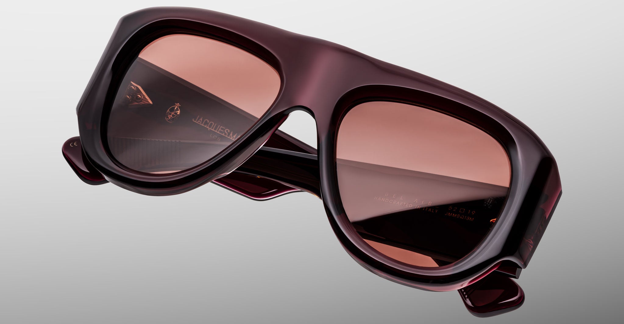 Close-up of Jacques Marie Mage Bel Air oversized sunglasses with thick, dark burgundy frames and tinted lenses on a light gray background. Subtle brand insignia and detailing appear inside the frame.