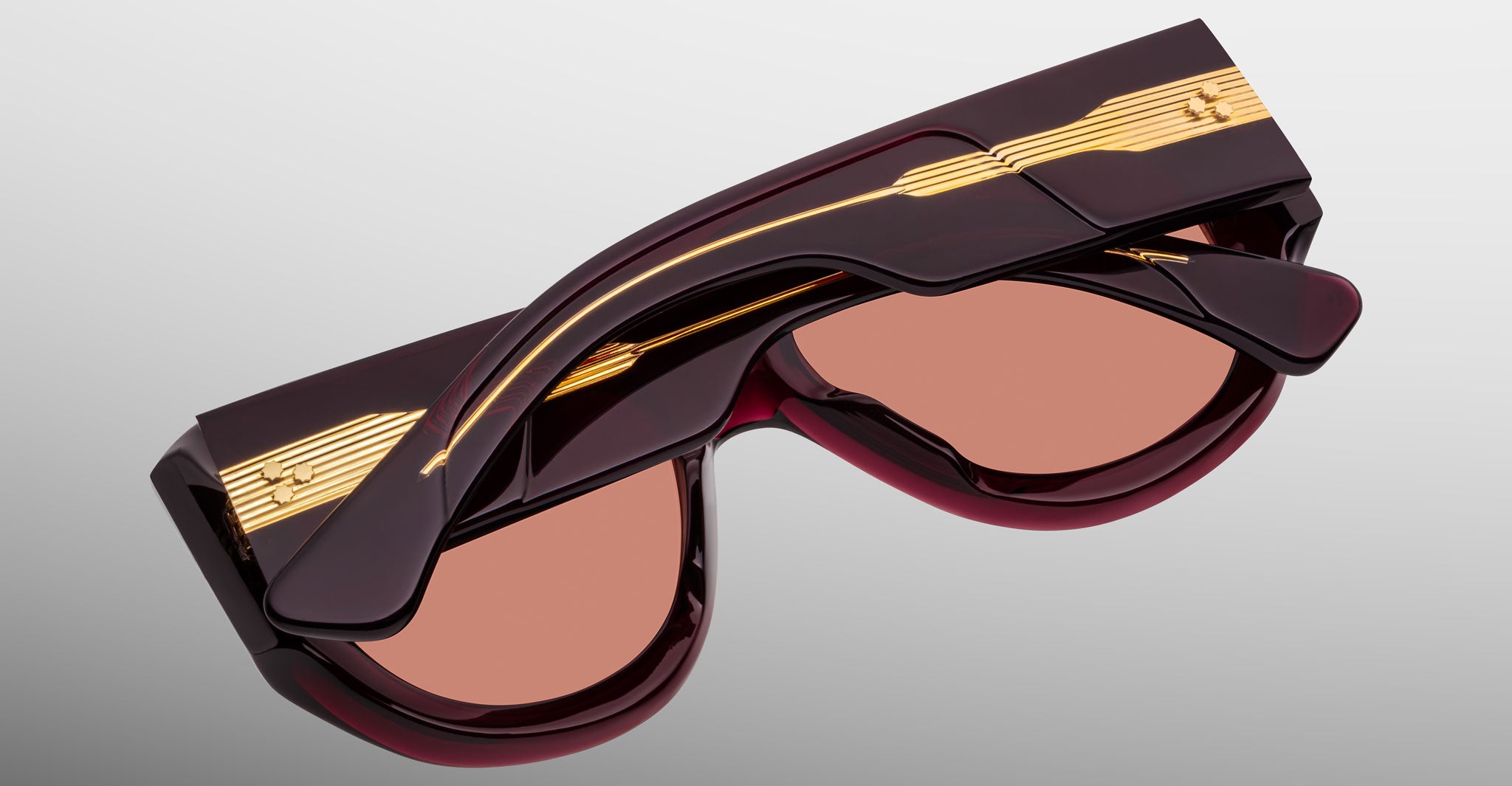 Jacques Marie Mage Bel Air sunglasses with dark maroon frames, gold temple accents, and pink-tinted lenses are displayed folded on a light gray background.