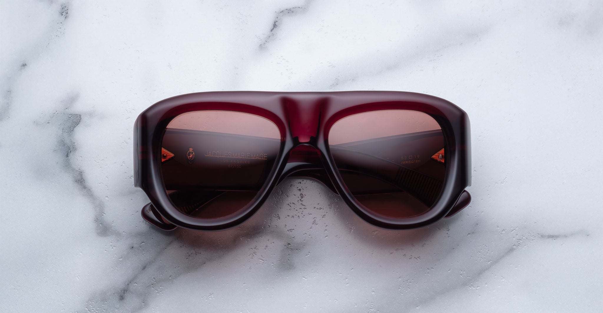 Jacques Marie Mage Bel Air sunglasses, featuring large dark red frames and tinted lenses, are displayed on a white marble surface. The brand name appears subtly on the inside of one arm.