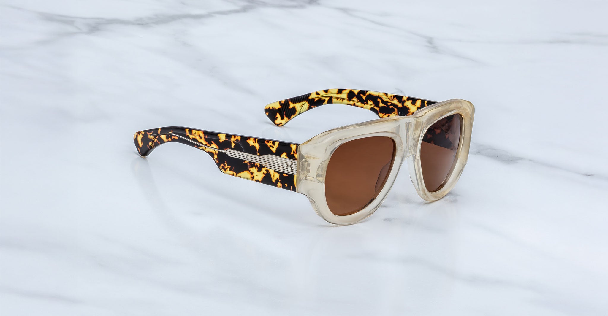 A pair of Jacques Marie Mage Bel Air pilot sunglasses with thick, transparent beige handcrafted acetate frames, tortoiseshell-patterned arms, and brown lenses rests on a white marble surface.
