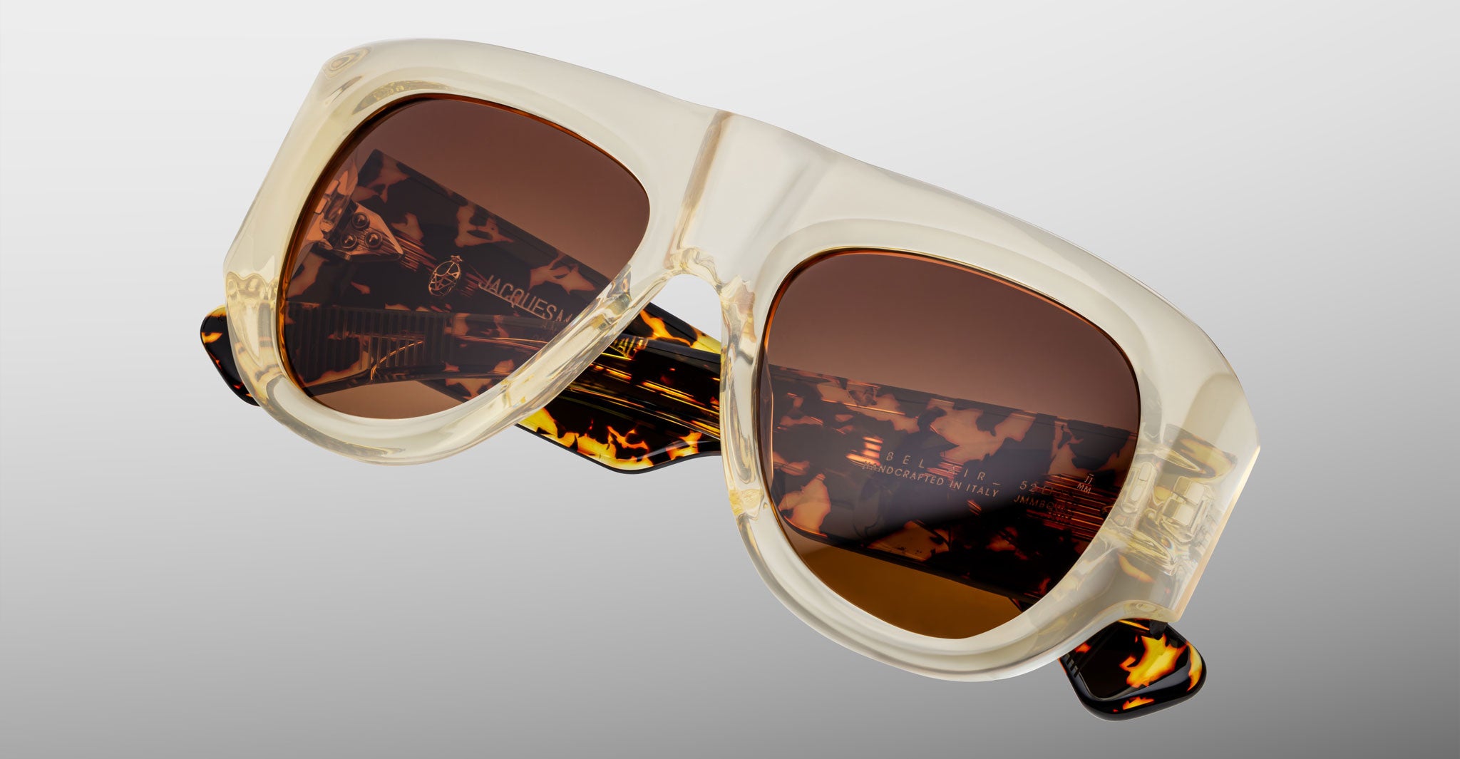 Close-up of Jacques Marie Mage Bel Air sunglasses with handcrafted translucent beige acetate frames, brown-tinted lenses, and tortoiseshell-patterned arms on a light gray background.
