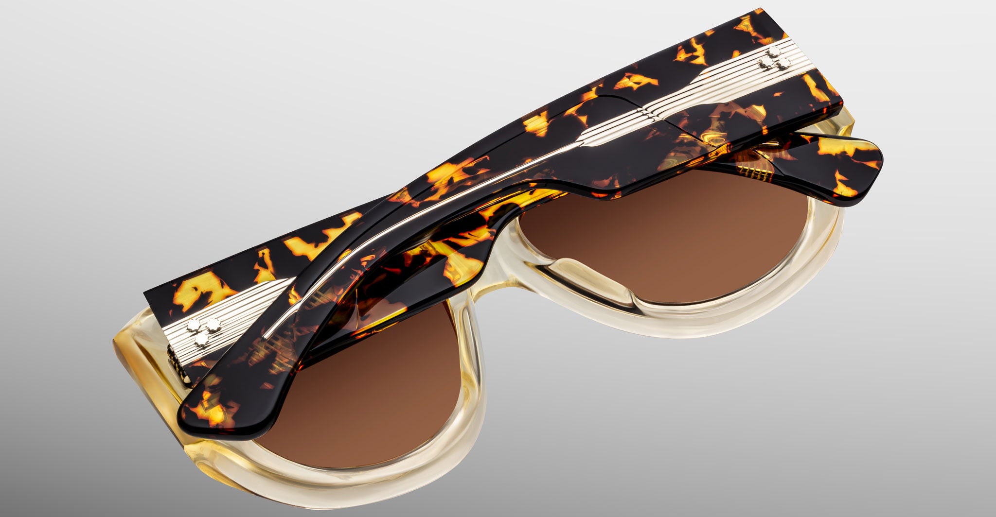 Close-up of Jacques Marie Mage Bel Air sunglasses featuring handcrafted acetate frames in a tortoiseshell pattern, metallic accents, and round brown lenses. The stylish folded arms rest atop a gradient gray background.