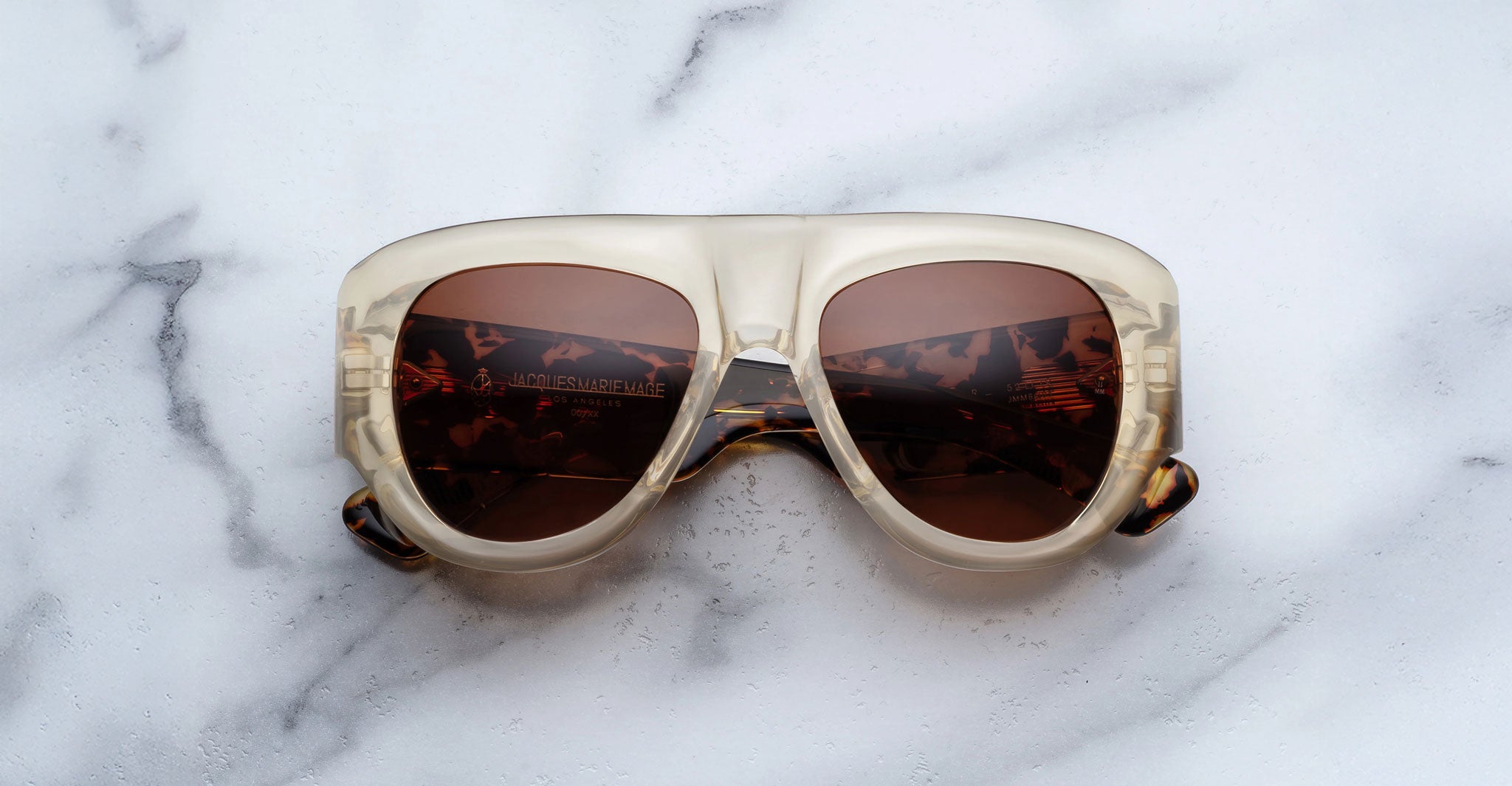 A pair of Jacques Marie Mage Bel Air sunglasses featuring oversized translucent beige handcrafted acetate frames and brown-tinted lenses sits on a white marble surface.