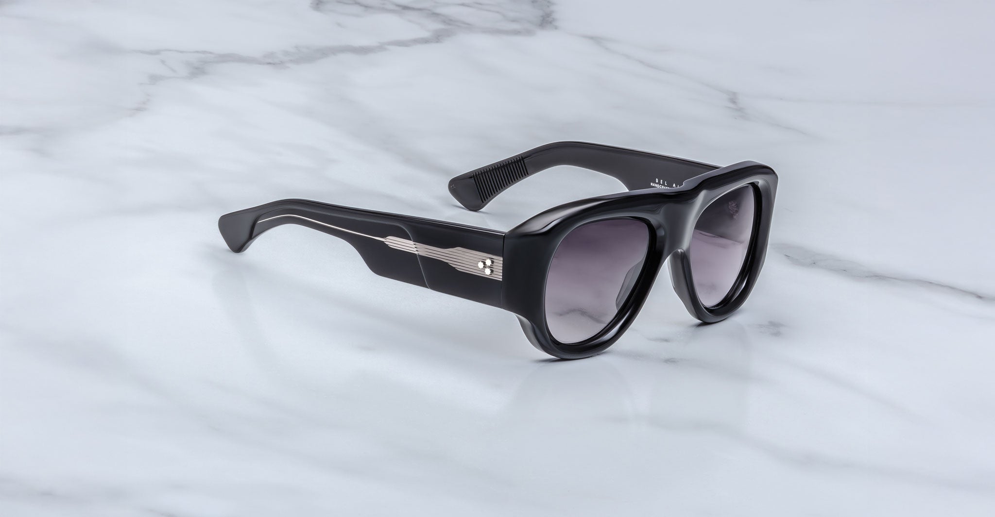 The Jacques Marie Mage Bel Air sunglasses feature thick black frames, dark lenses, and silver metallic temple details, stylishly displayed on a white marble surface.