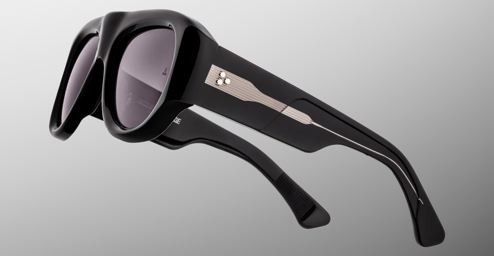 A close-up of the Jacques Marie Mage Bel Air sunglasses, showcasing thick black glossy frames, dark lenses, and temples adorned with a silver metallic accent featuring three studs and a linear design against a plain gradient background.