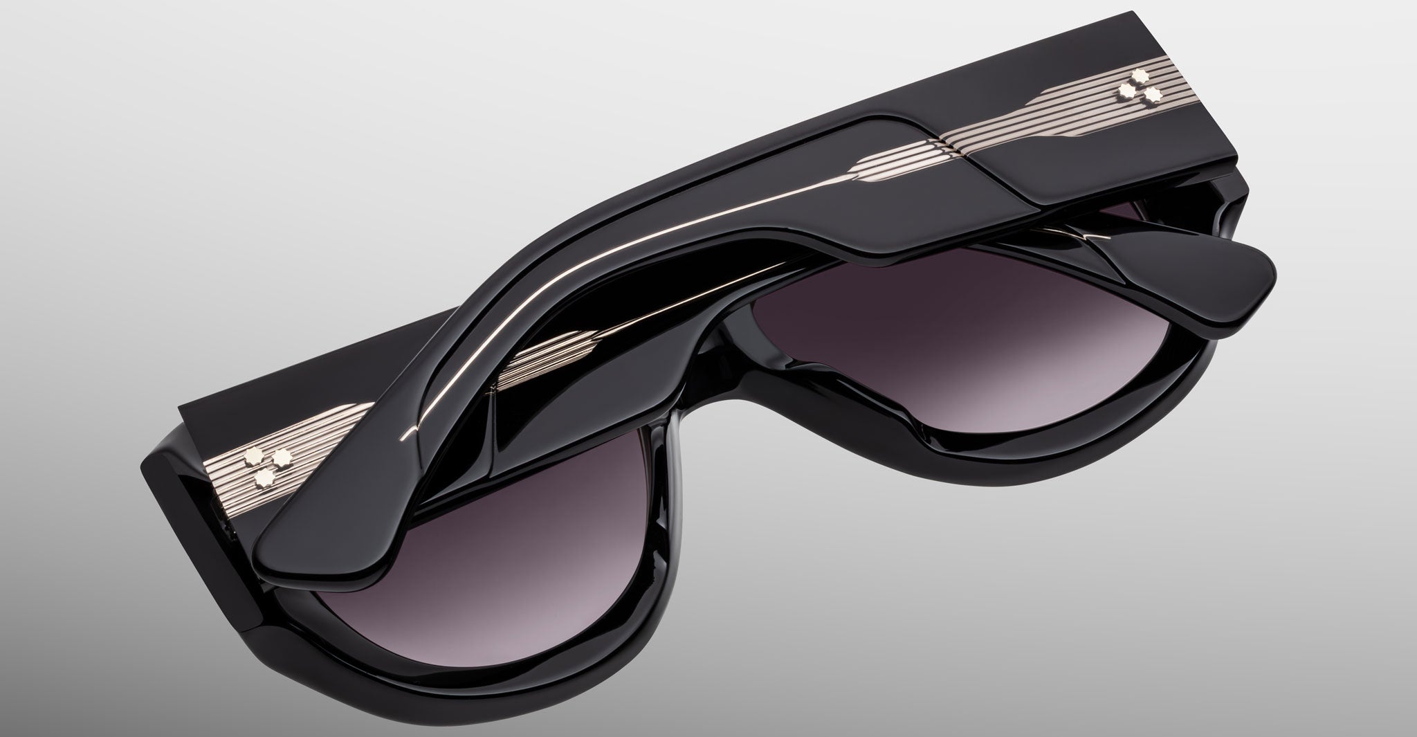 The Jacques Marie Mage Bel Air sunglasses in glossy black feature folded arms with metallic line designs and small dot accents on the temples, showcased against a light gray background.