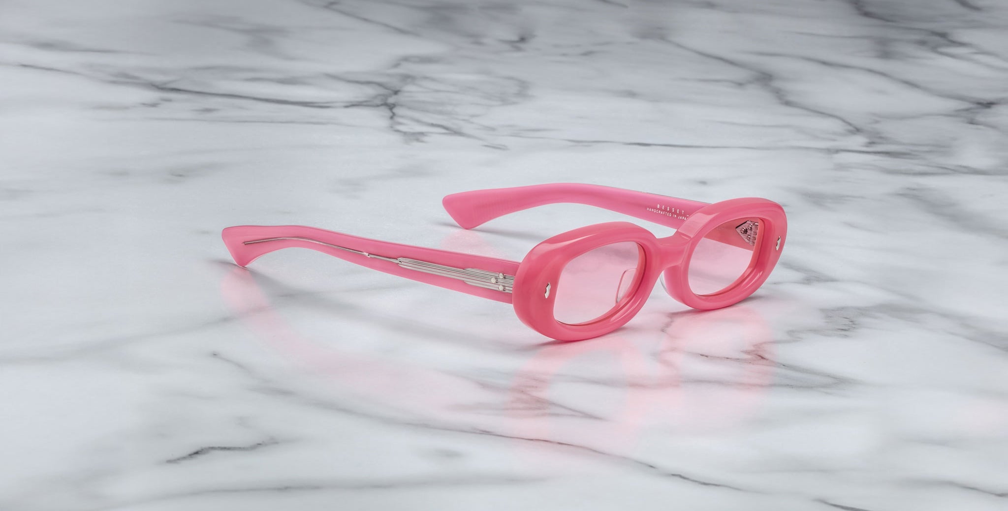 The Jacques Marie Mage Besset sunglasses, featuring round pink frames and tinted lenses, rest on a white marble surface with grey veining.
