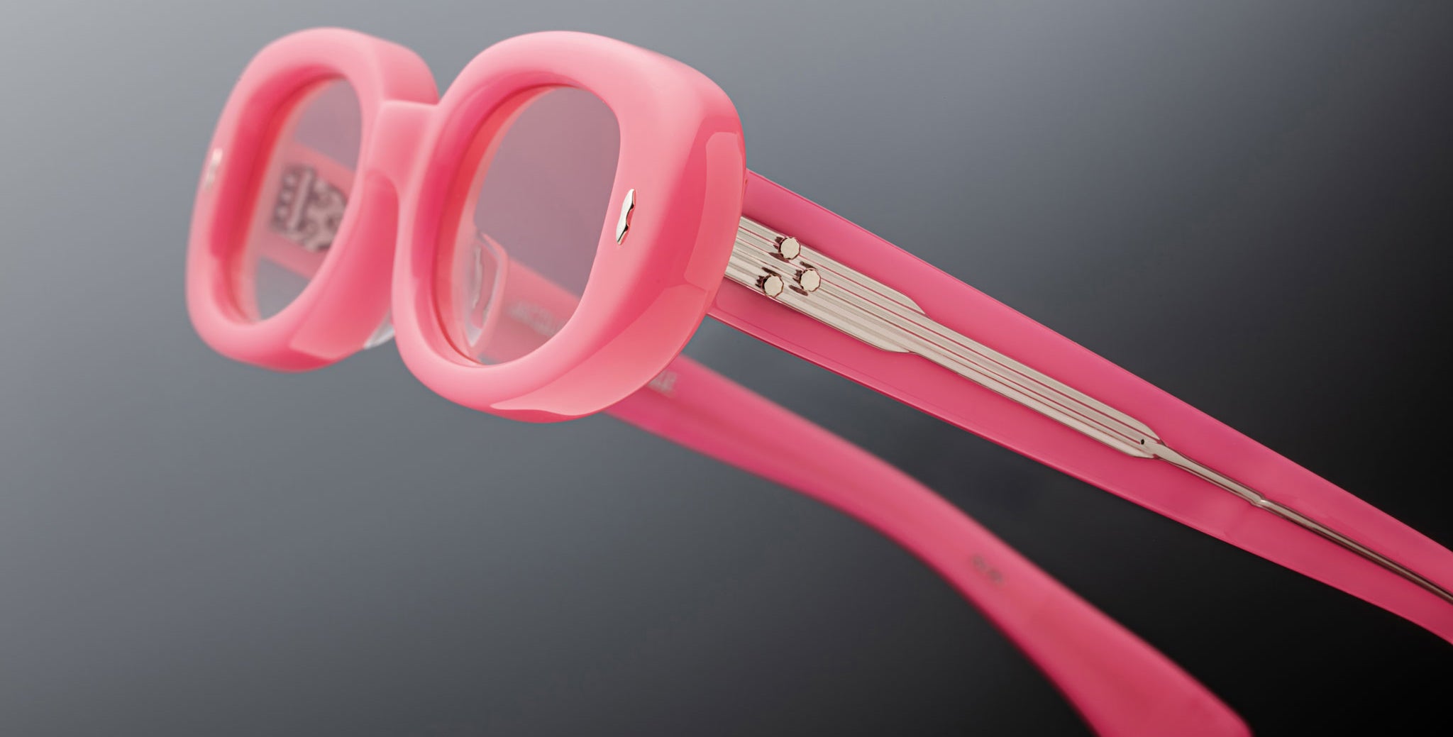 Close-up of Jacques Marie Mage Besset eyeglasses featuring thick, rounded pink frames and matching temples with visible metal hinges adorned with a linear design. The background is a soft, blurred gray.