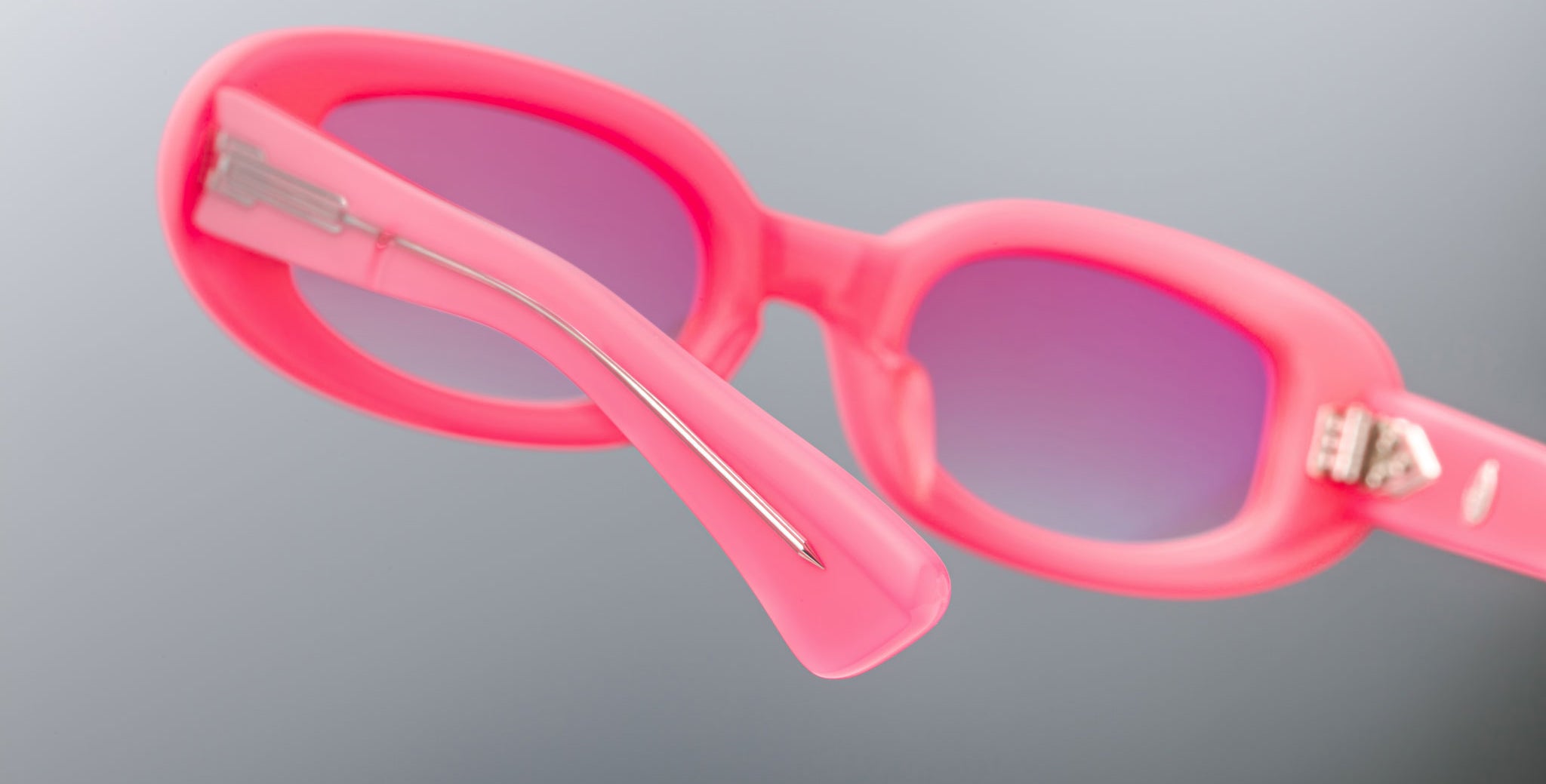 Close-up of Jacques Marie Mage Besset pink sunglasses with gradient purple lenses, highlighting the inside of the frame, arm, and visible metal hinge and bar against a plain gray background.