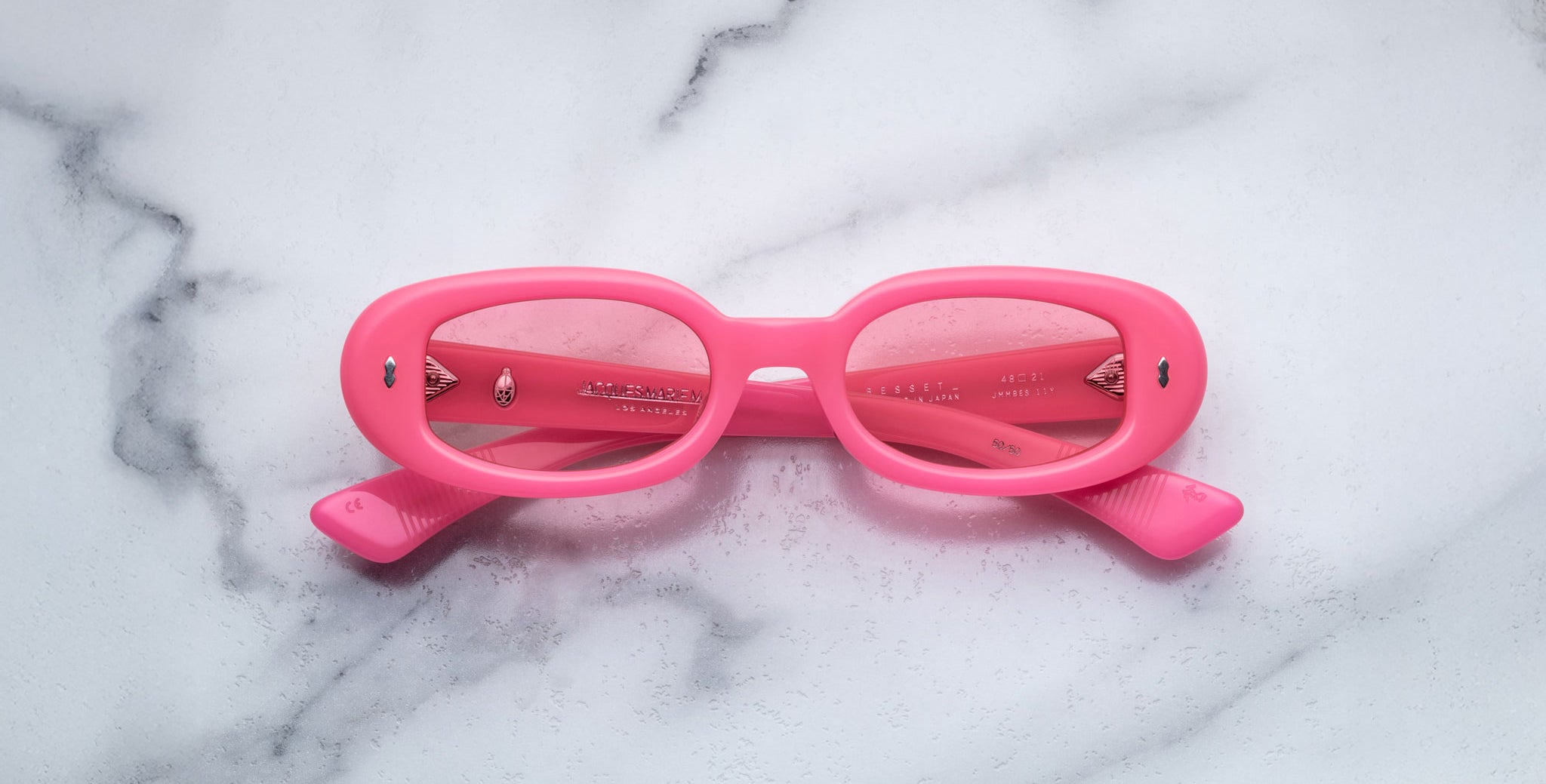 A pair of Jacques Marie Mage Besset bright pink oval sunglasses with tinted lenses is placed upside down on a white marble surface.