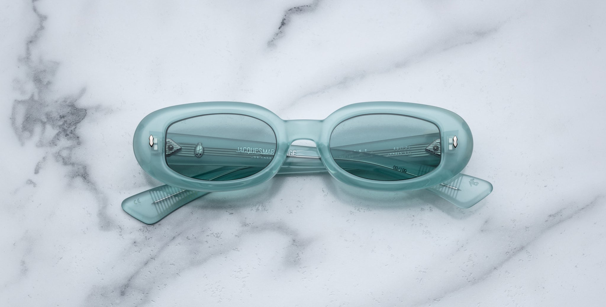The Jacques Marie Mage Besset sunglasses, light blue and oval-shaped with tinted lenses, rest on a white marble surface with their arms slightly open and visible.