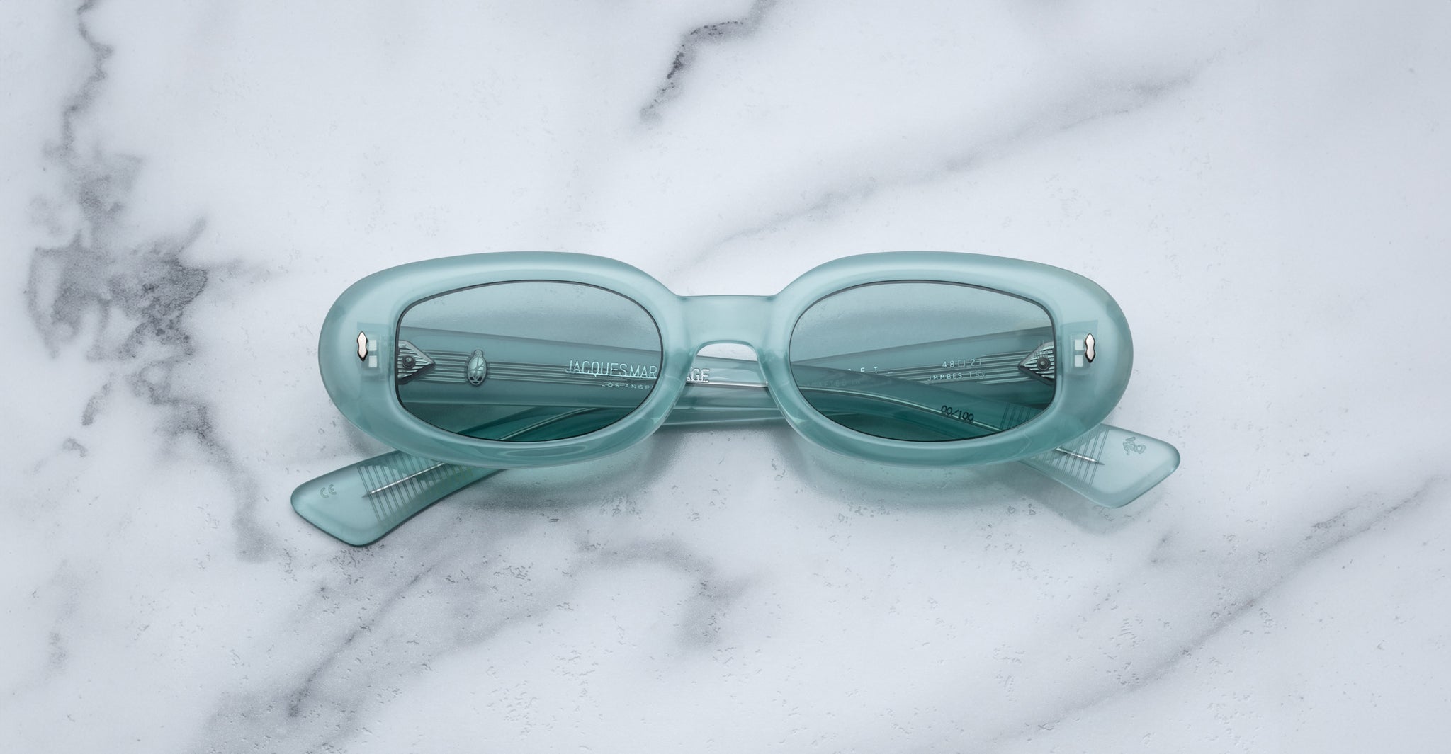 The Jacques Marie Mage Besset sunglasses, light blue and oval-shaped with tinted lenses, rest on a white marble surface with their arms slightly open and visible.
