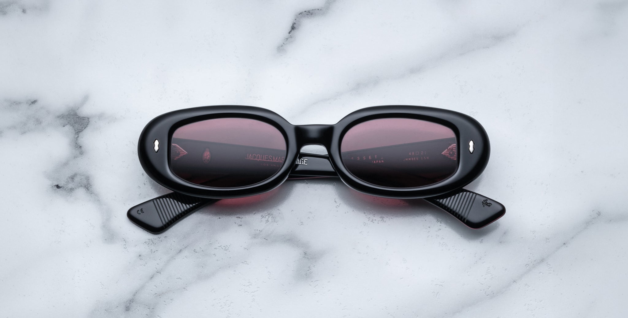 A pair of Jacques Marie Mage Besset black oval sunglasses with dark lenses rests face-up on a white marble surface with gray veining, their arms crossed in an X shape.