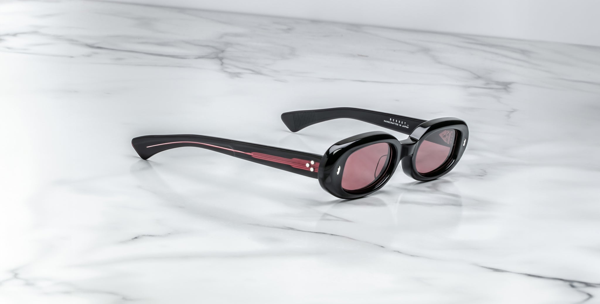 Jacques Marie Mage Besset black oval sunglasses with pink-tinted UV lenses, shown from above.