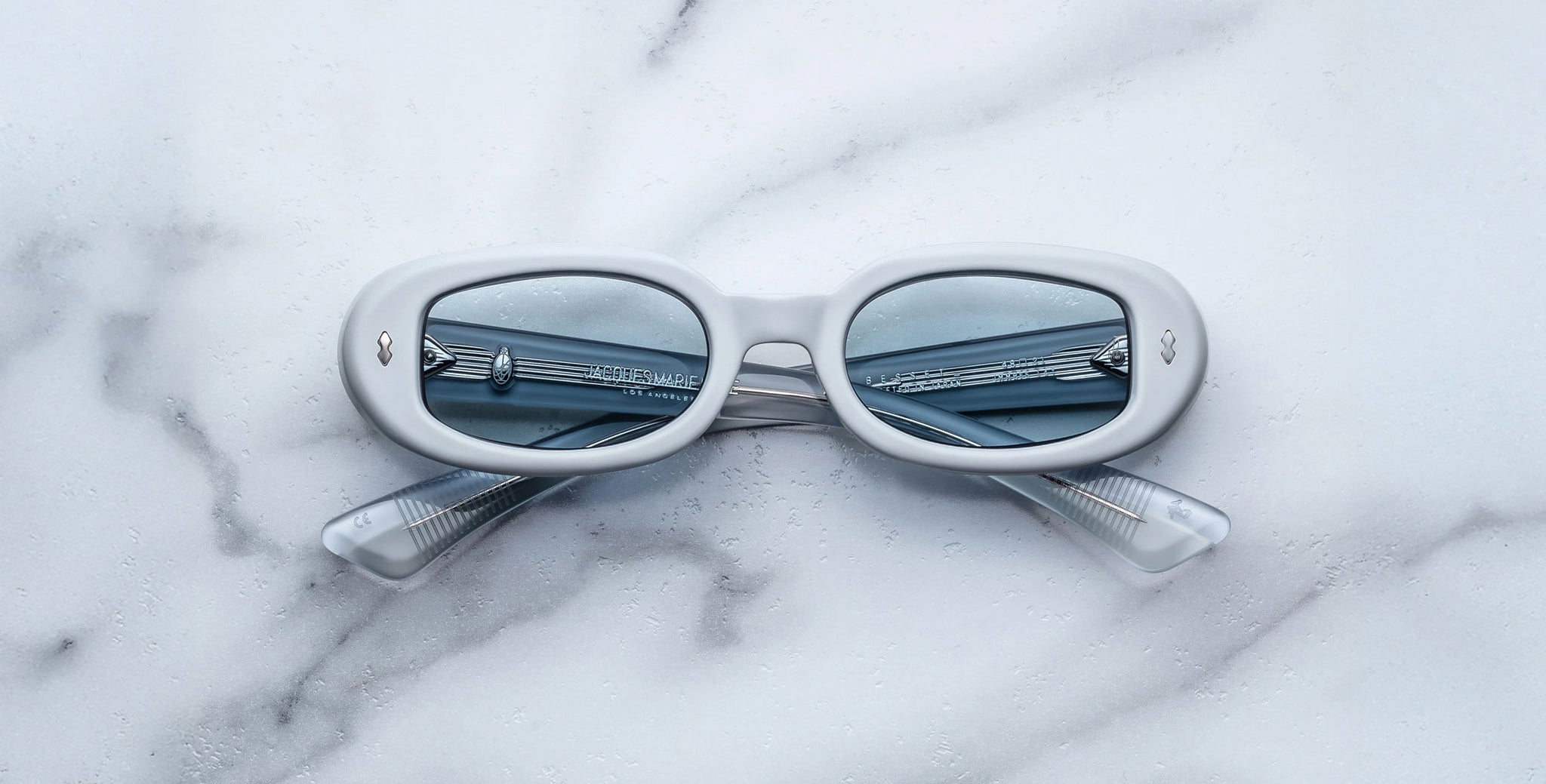 A pair of Jacques Marie Mage Besset white oval sunglasses with blue-tinted lenses rests on a white marble surface. The crossed temples and a metal railing reflected in the lenses add to their striking appearance.