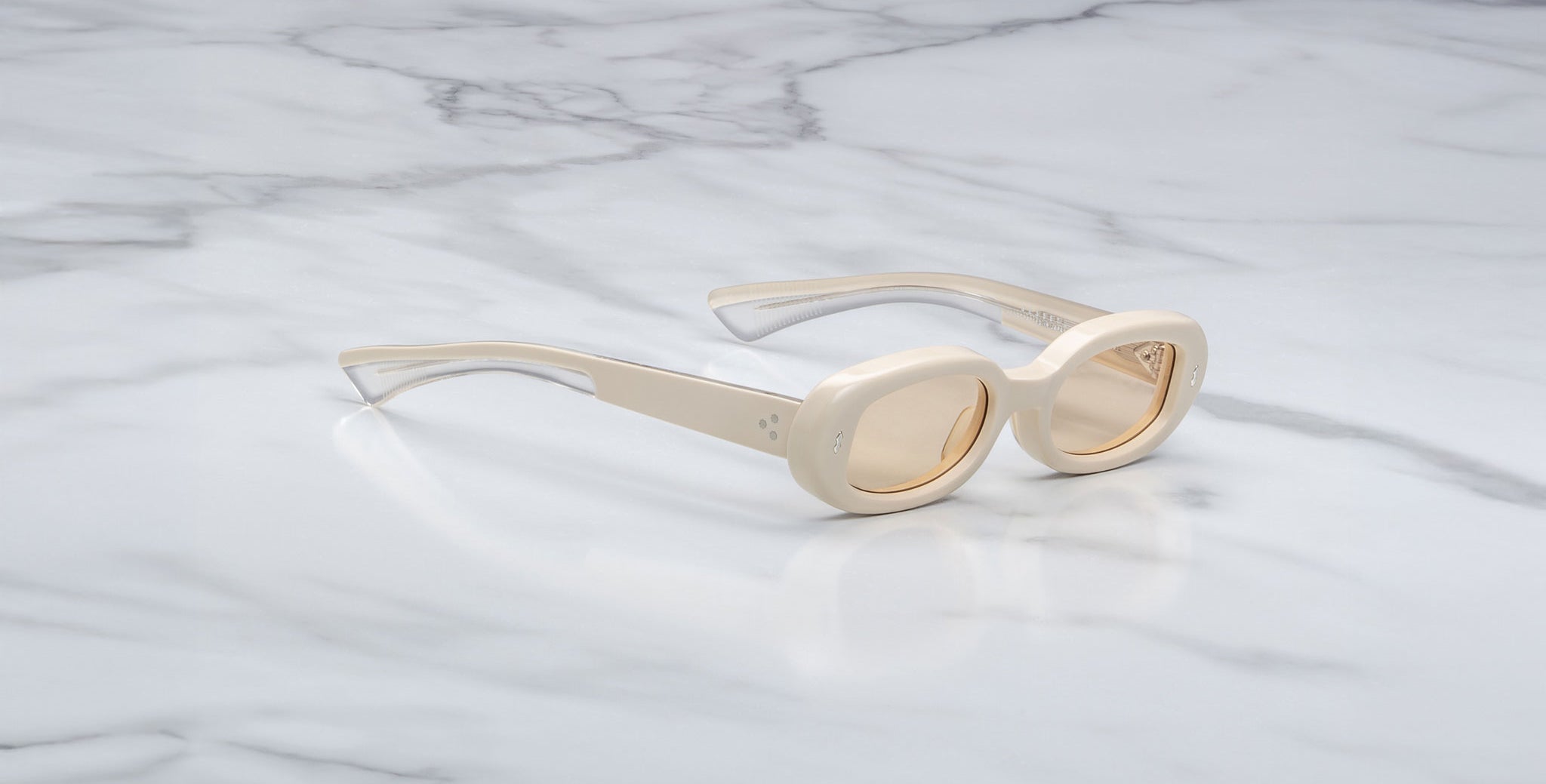 A pair of Jacques Marie Mage Besset cream-colored, oval sunglasses with light brown tinted lenses rests on a white marble surface with gray veining.
