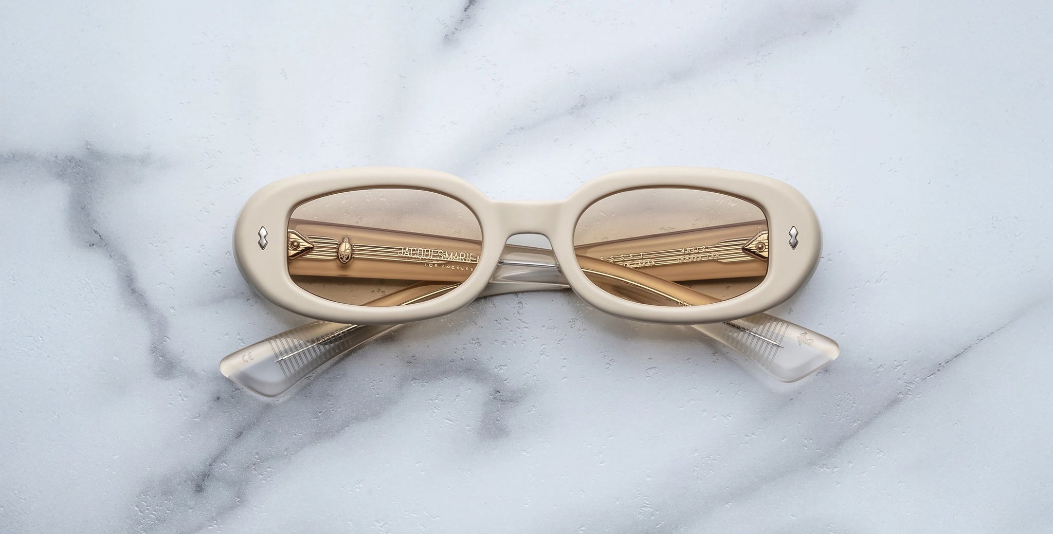 The Jacques Marie Mage Besset, a pair of cream-colored oval sunglasses with tinted lenses and folded arms, rests upside down on a white marble surface.
