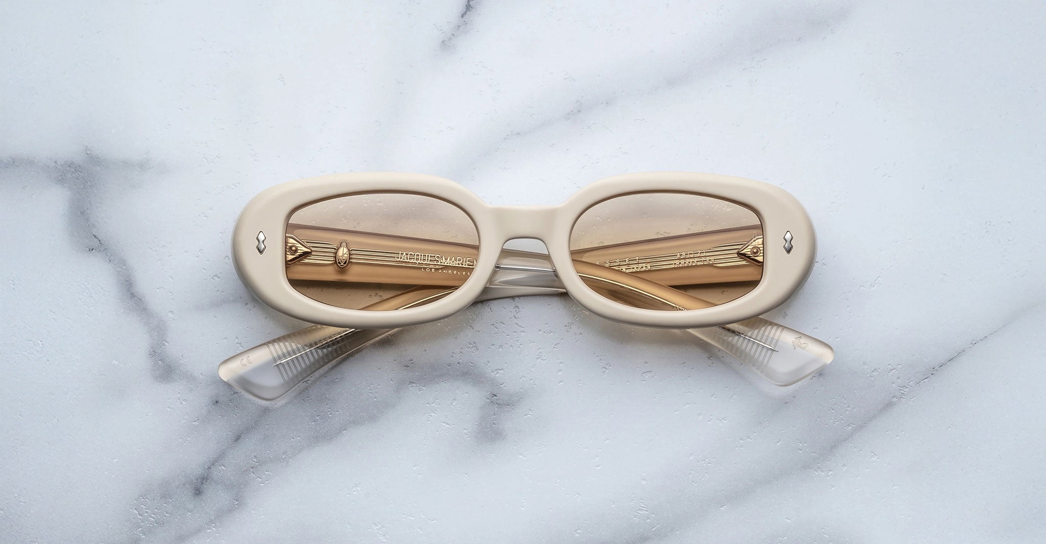 The Jacques Marie Mage Besset, a pair of cream-colored oval sunglasses with tinted lenses and folded arms, rests upside down on a white marble surface.