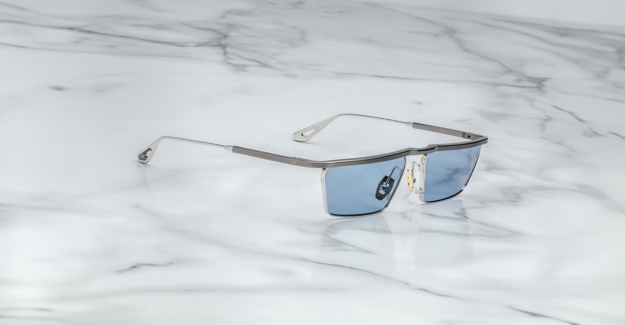 The Jacques Marie Mage Bogart features modern, rectangular blue lenses and thin silver frames, displayed on a white marble surface with gray veining.