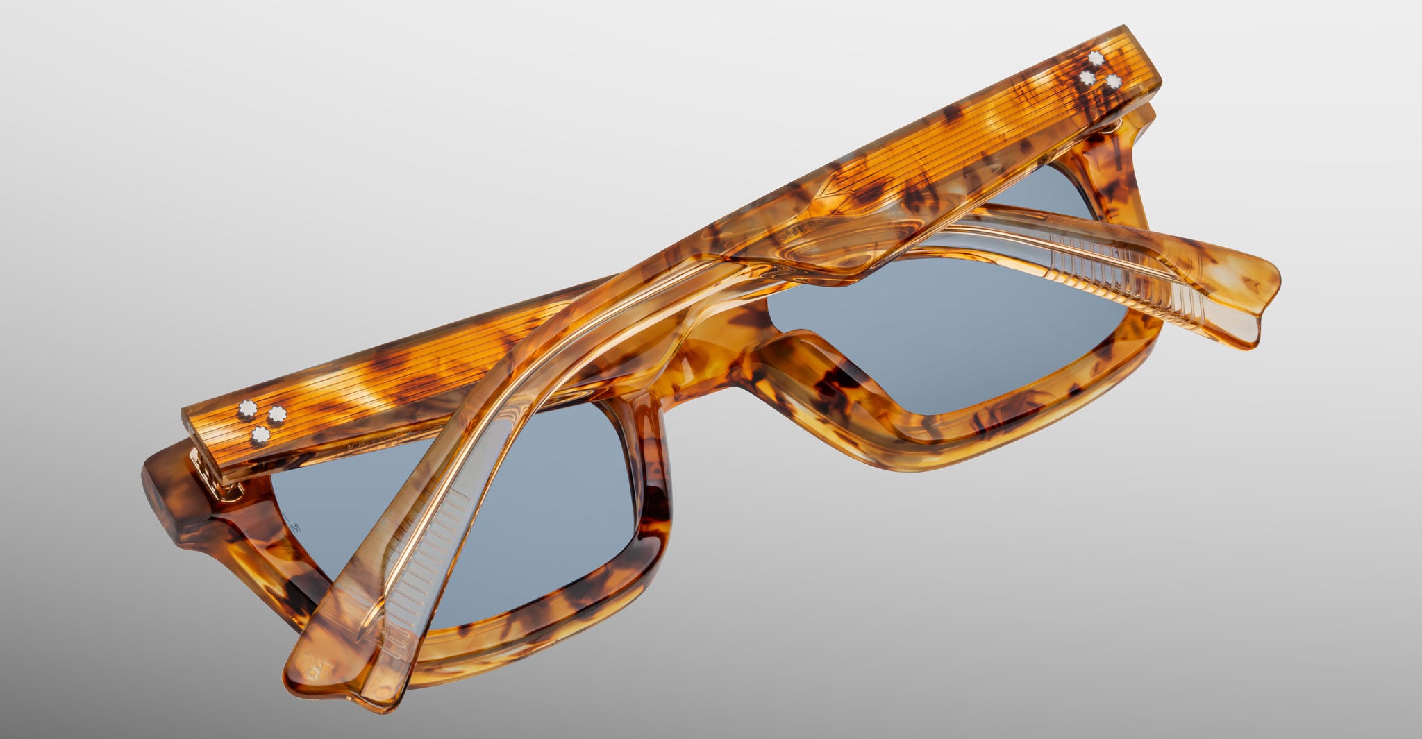 The Jacques Marie Mage Casablanca sunglasses, featuring a marbled brown tortoiseshell frame with blue lenses and silver accents on the front corners, are shown folded on a light grey surface.