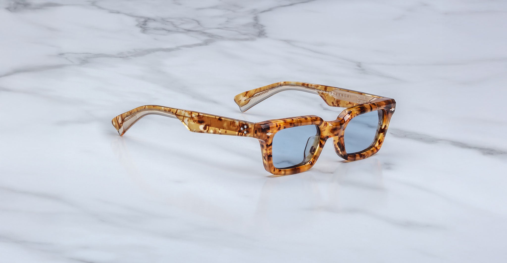 The Jacques Marie Mage Casablanca sunglasses feature thick, amber and brown tortoiseshell rectangular frames with blue-tinted lenses, displayed on a white marble surface.